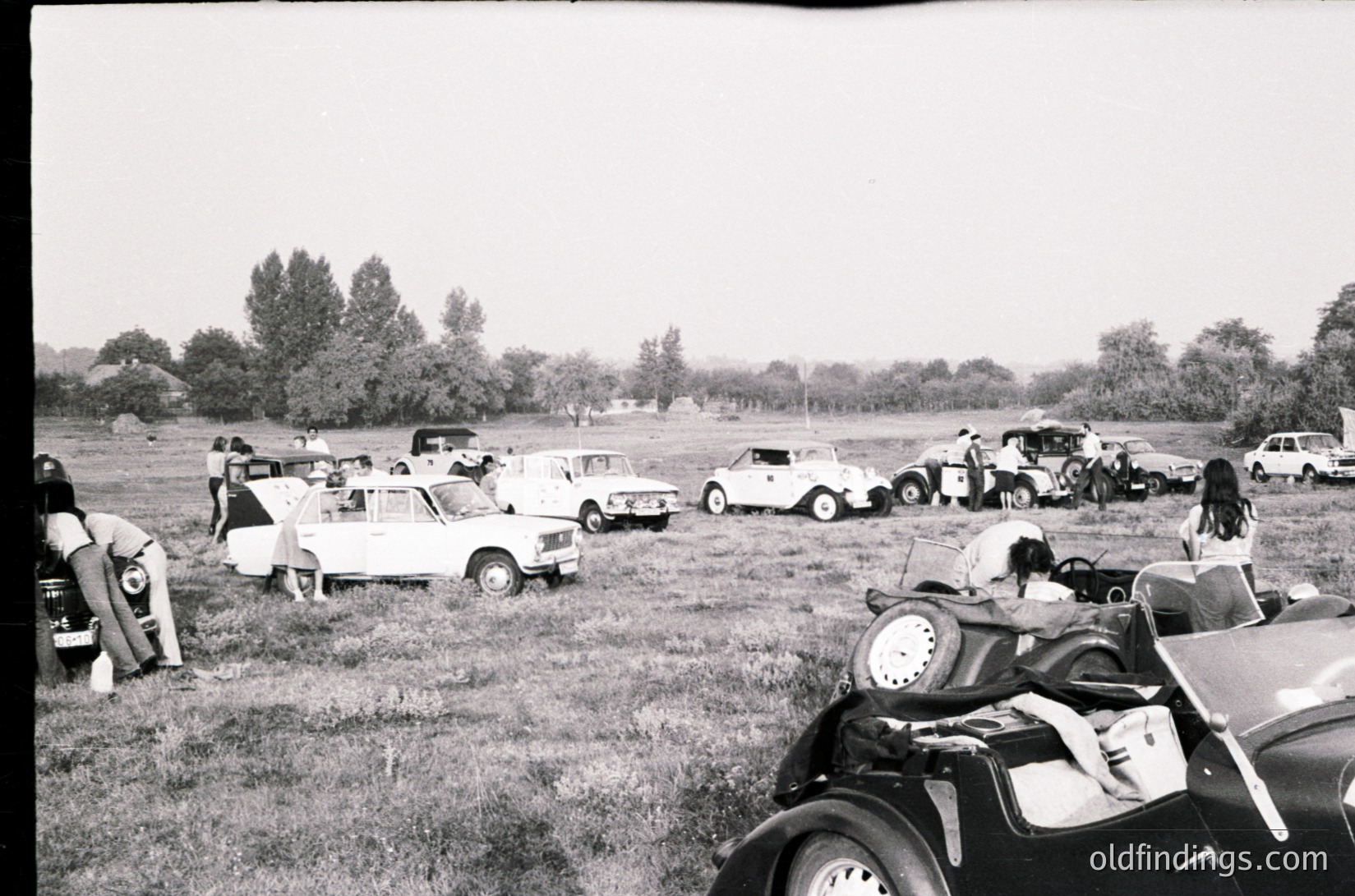 Vintage car rally in open field with mid-century compact cars and classic vehicles, likely 1960s–1970s. Participants inspect engines or chat near parked cars, suggesting a gathering or meetup. Rustic, rural setting with scattered trees and overcast sky.