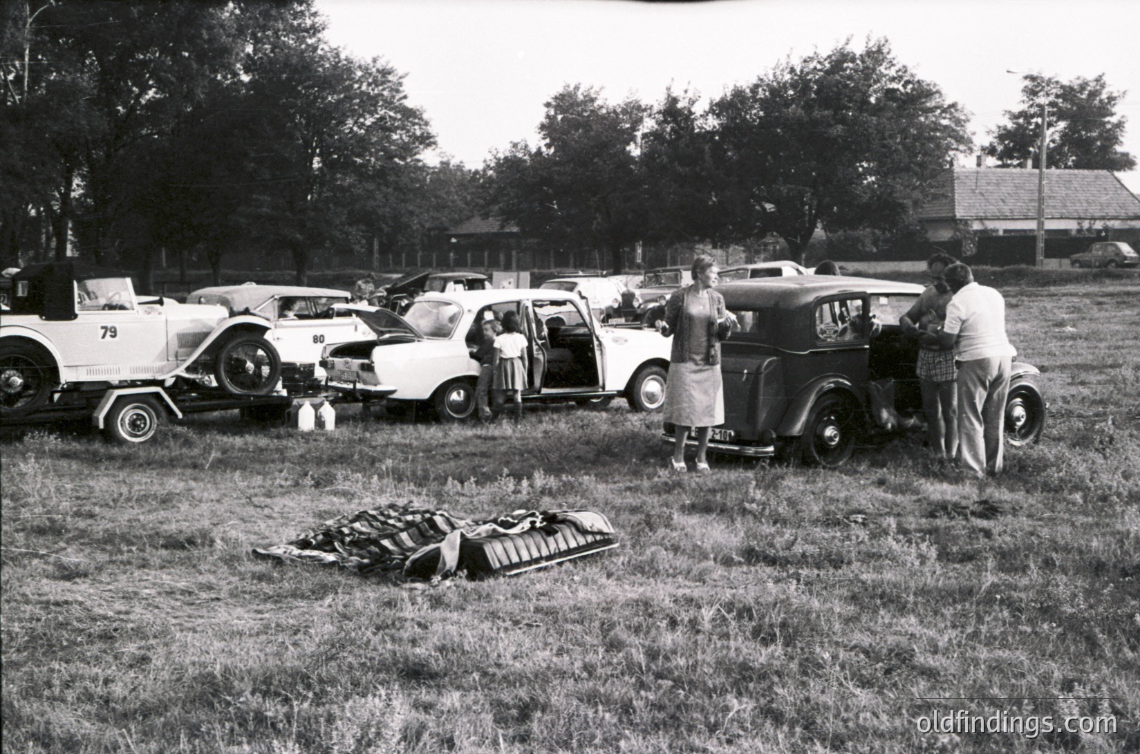 Vintage outdoor gathering featuring classic cars (1950s–1960s) parked on grass, including a trailer, station wagon, and sedan. Group of people inspecting vehicles, with one man standing near an open trunk holding a folded blanket. Rustic rural setting with trees and a distant building.