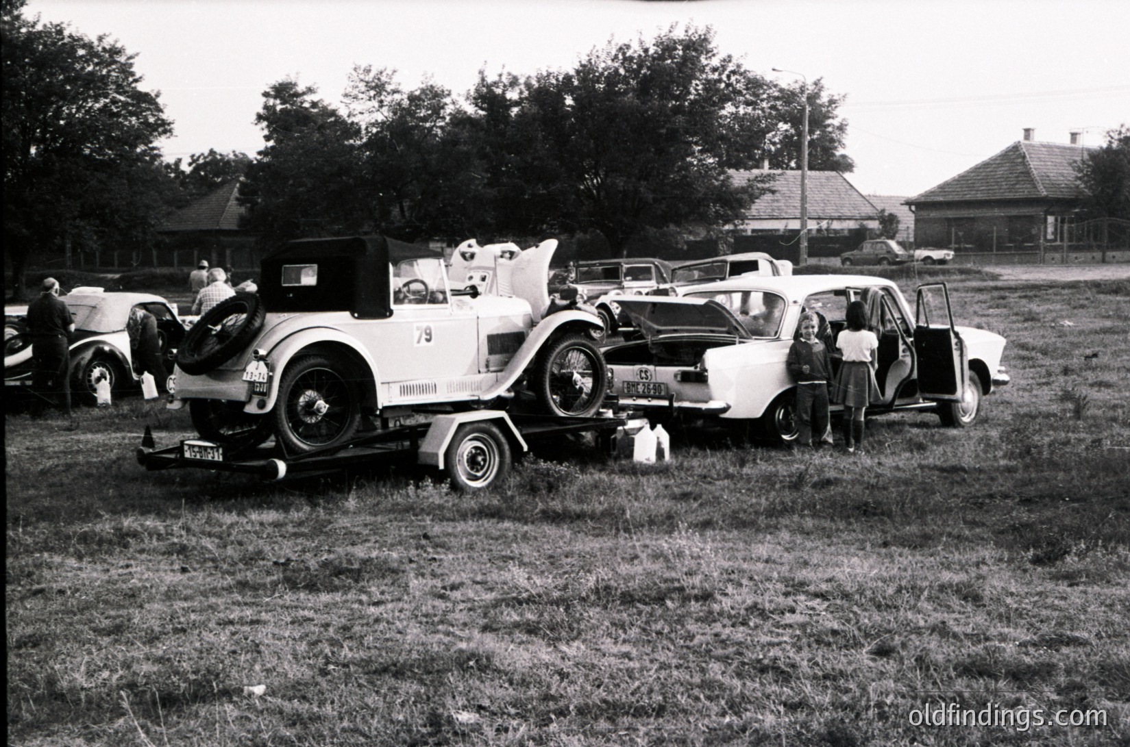 Vintage car show featuring classic automobiles, likely mid-20th century (1940s–1950s). Three open-top vehicles dominate: a white roadster with "Mazda" badge, a cream-colored sedan, and a smaller vintage car. Group of people in casual attire inspects or poses near vehicles. Rural setting with grassy field, trees, and a distant building. Ideal for automotive history, nostalgia, or stock photography.