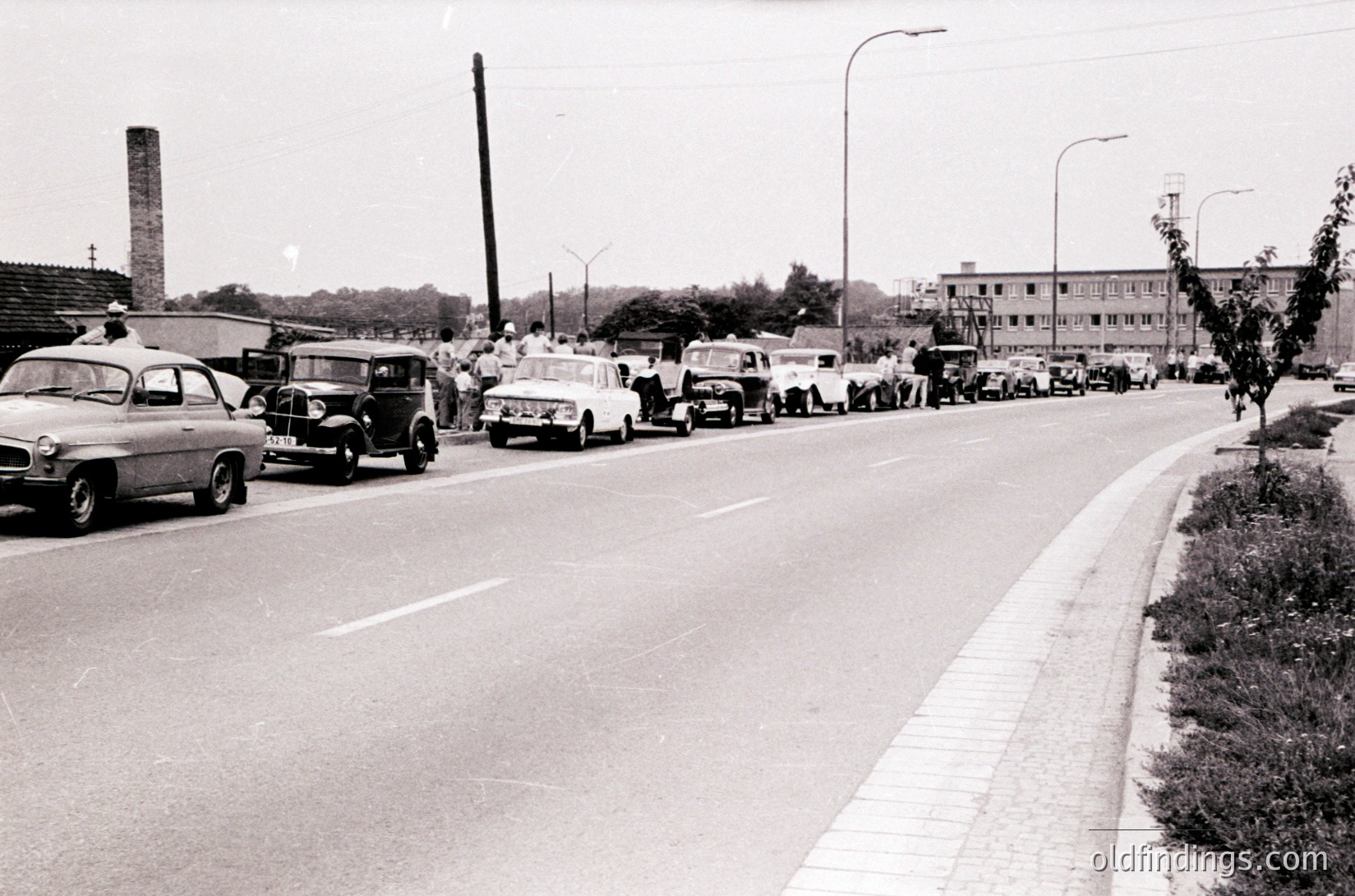 Mid-20th century urban street jammed with vintage cars, likely 1950s–1960s. Dense traffic of classic sedans, station wagons, and a military truck on a wide, tree-lined road. Industrial chimney and mid-rise apartment buildings in background suggest post-war European city.