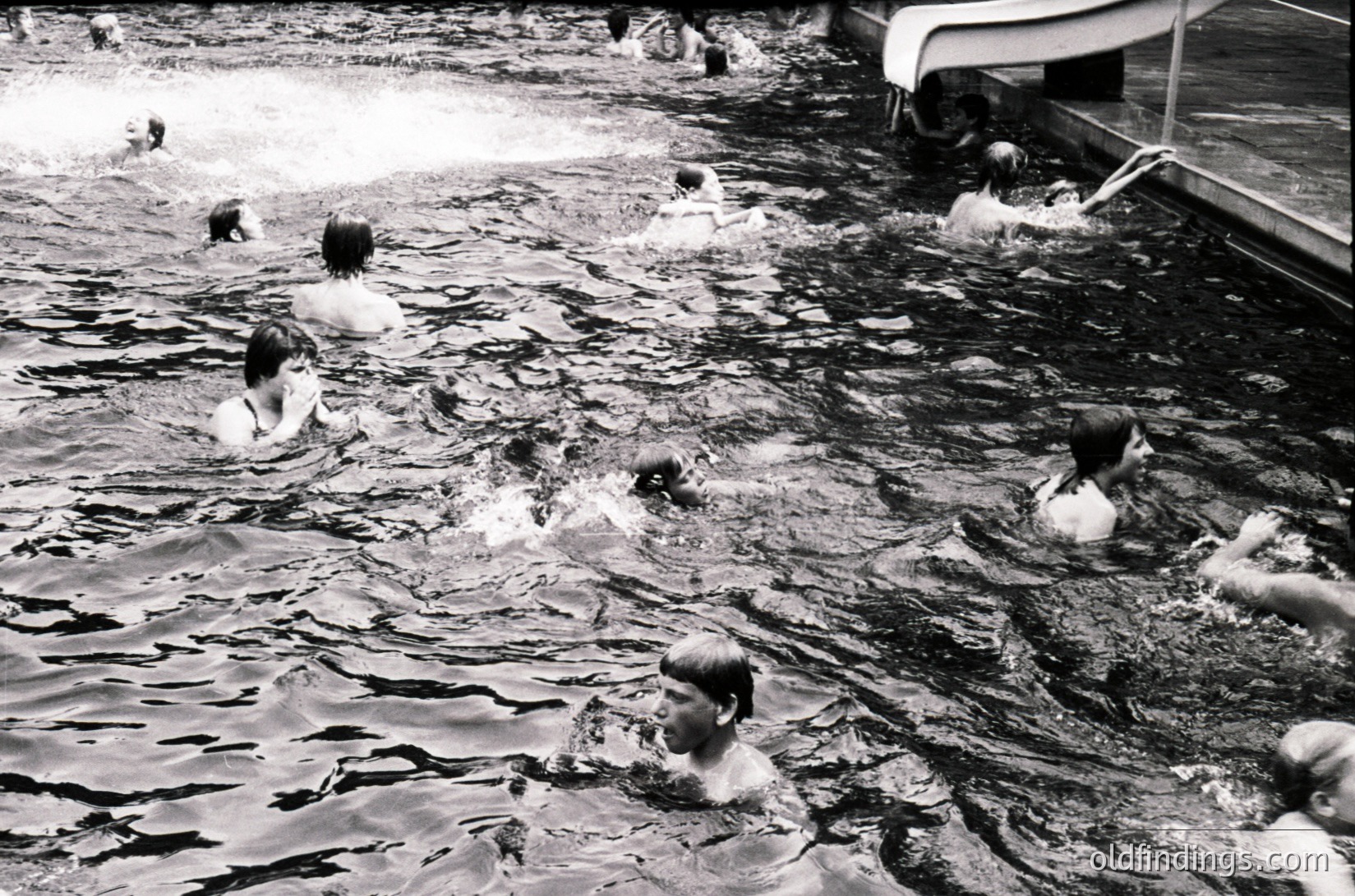 Vintage black-and-white photo of a lively seaside scene with children playing in shallow waves, mid-20th century. Wooden pier and boat dock in background suggest a coastal resort. Casual swimwear reflects mid-century beach culture.