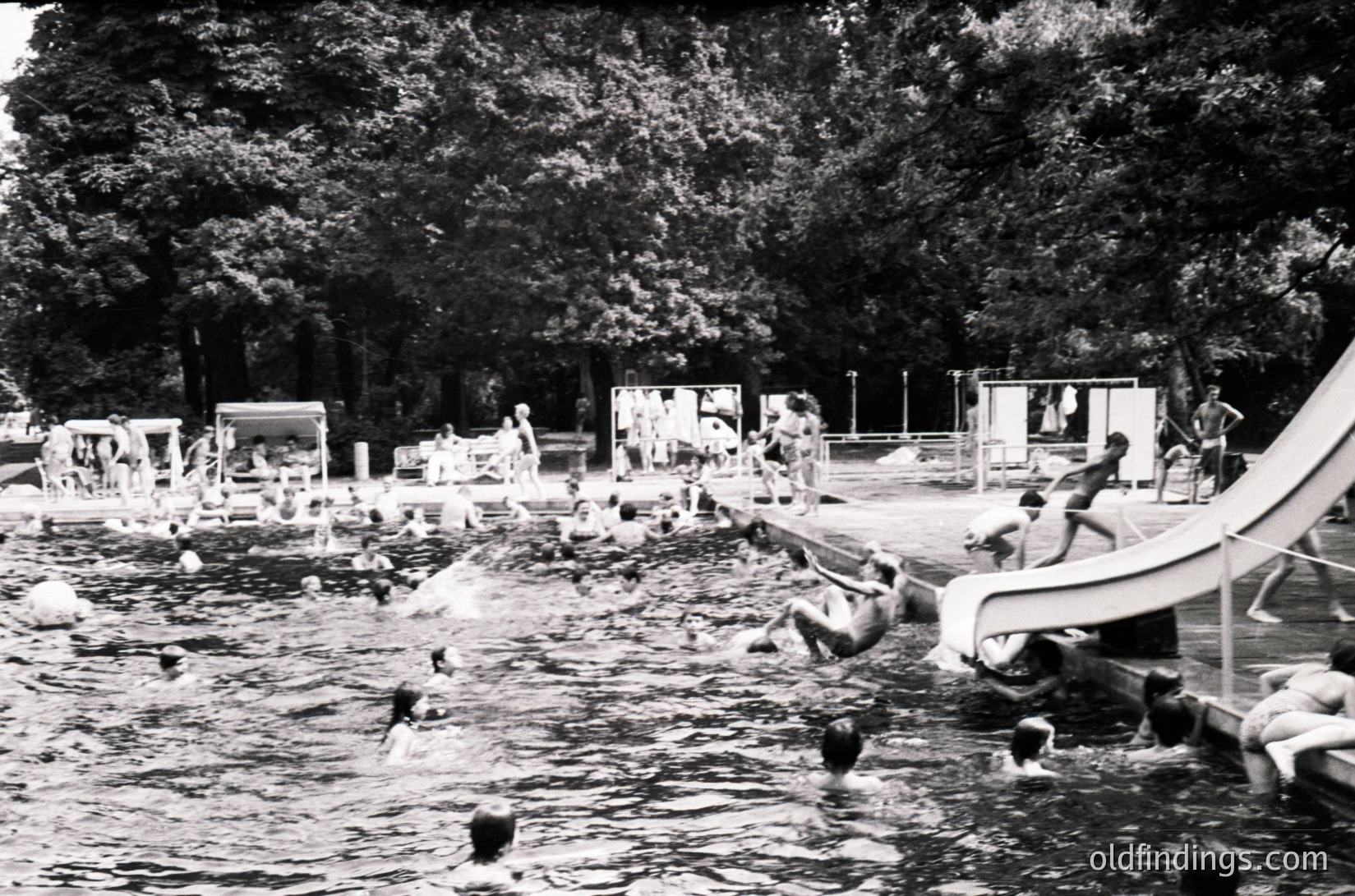 Vintage black-and-white photo of a mid-20th-century outdoor swimming pool with a waterslide, surrounded by changing cabins and lush trees. Crowded with swimmers and children playing in shallow and deep areas. Reflects mid-century recreational culture.