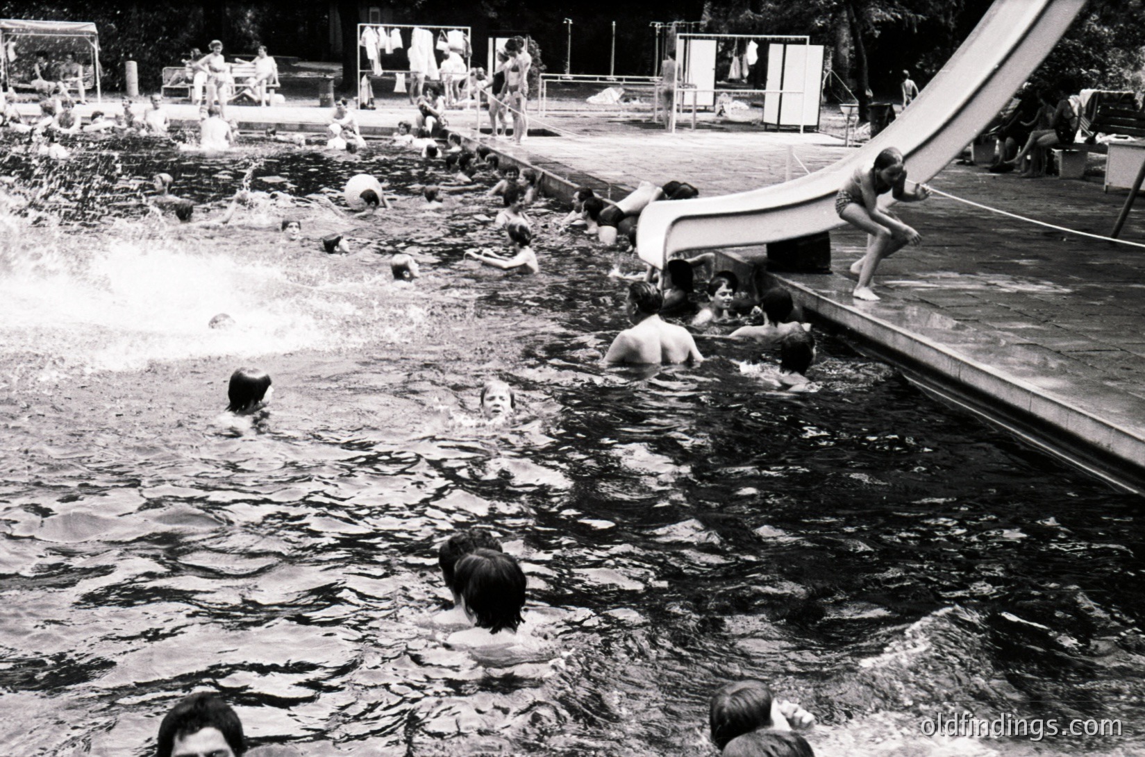 Vintage black-and-white shot of a lively outdoor pool with curved concrete edges and a diving platform. Crowded with swimmers, including children and adults, enjoying mid-century recreational culture. Surrounding area features trees and a paved promenade.