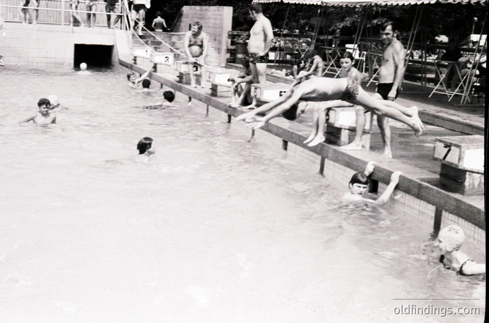 Vintage black-and-white shot of a mid-20th century outdoor swimming pool with diving board. Crowded with swimmers, children, and adults enjoying leisure time. Concrete pool with ladder entry, wooden diving platform, and adjacent changing area with umbrellas. Mid-century recreational architecture.