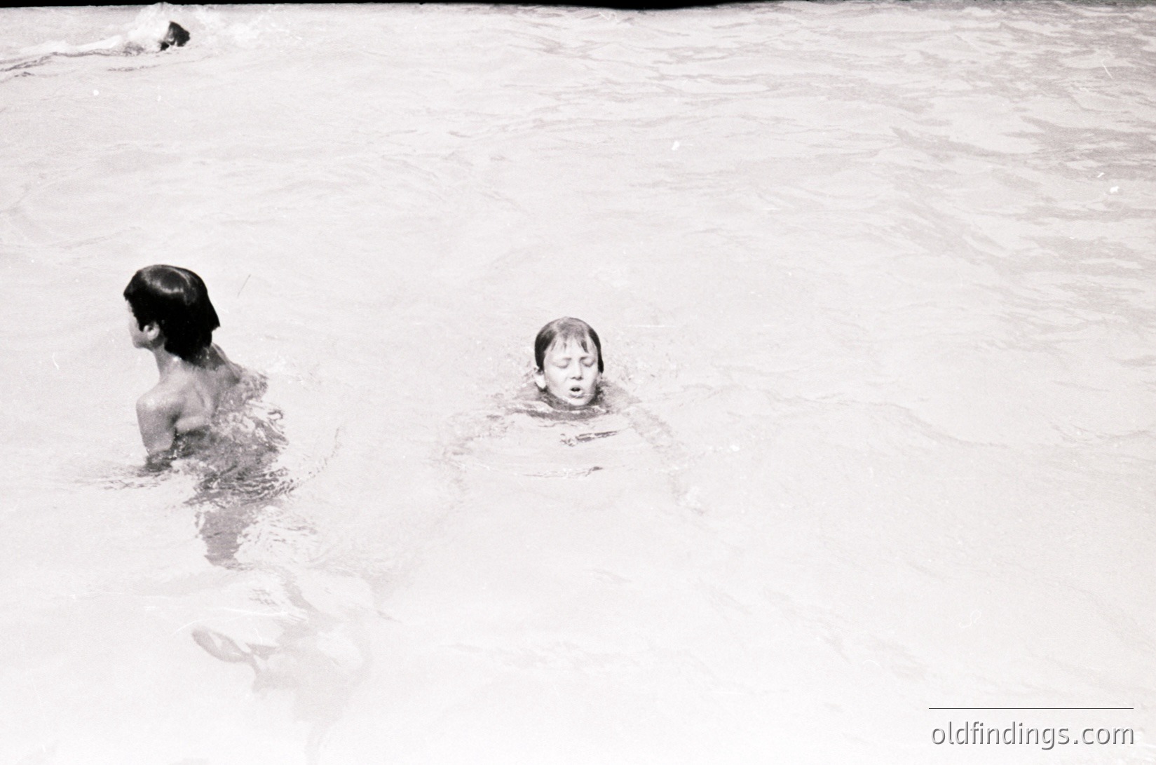 Black-and-white shot of two children swimming in shallow water, likely an indoor pool. The older child (left) appears to be teaching the younger (right) to float. Mid-20th century style swimwear and minimalist composition suggest a midcentury aesthetic.