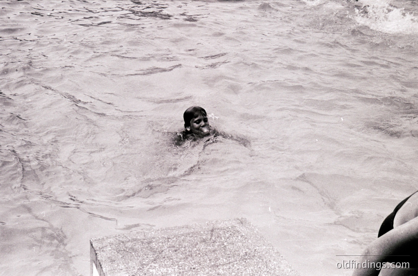 Black-and-white shot of a child swimming in shallow ocean waves, partially submerged with only head visible. Waves gently lap around them, suggesting calm seas. Partial view of a concrete structure (likely a pier or jetty) at bottom left.