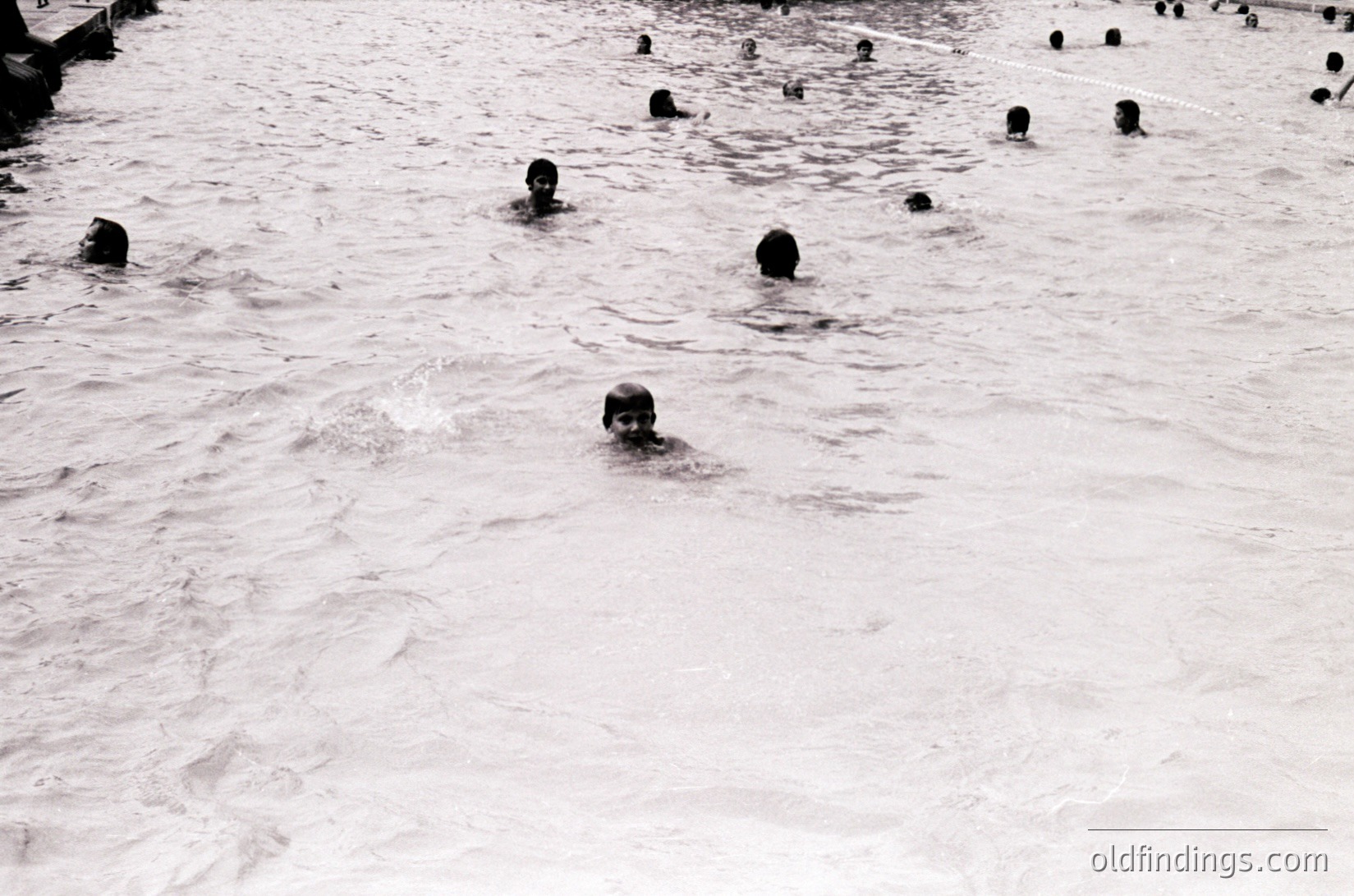 Black-and-white shot of a crowded outdoor swimming pool with numerous swimmers in shallow water, likely mid-20th century. Uniform swimwear suggests a public or school setting.