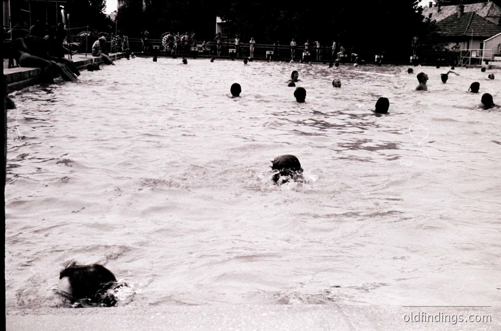 Black-and-white shot of a crowded outdoor swimming pool, likely from the 1960s–1970s. Numerous swimmers, including children and adults, engage in playful activity amid churned water. Fenced perimeter and distant trees suggest a public or resort setting. Ideal for historical research on leisure trends.