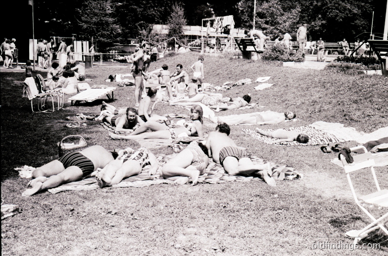 Mid-century beach scene with sunbathers lounging on towels and deck chairs in a grassy, open-air setting. Wooden structures and picnic tables suggest a resort or public park. Black-and-white style indicates 1950s–1970s era. Casual summer attire reflects mid-20th-century leisure culture.