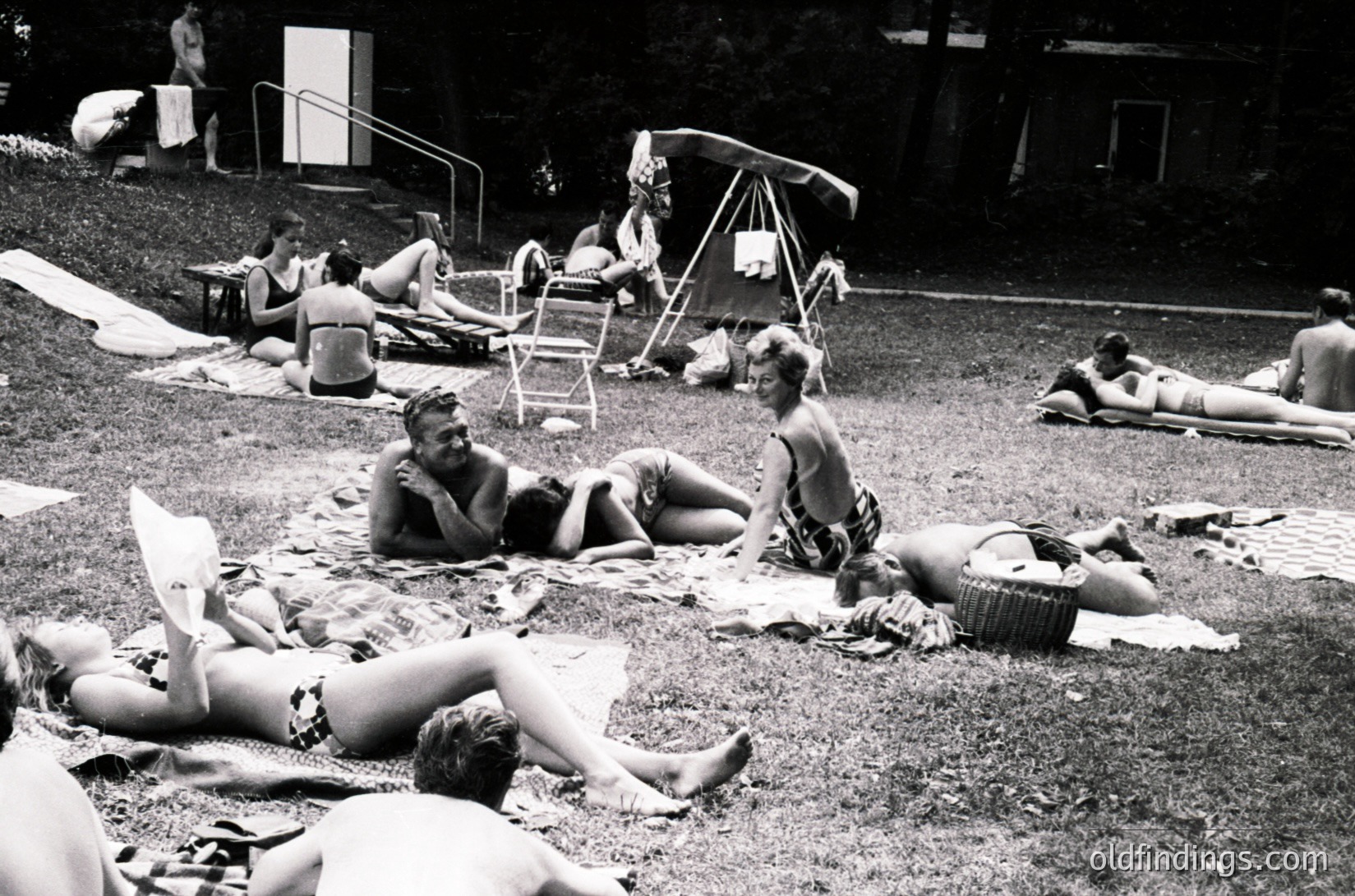 Vintage black-and-white beach scene from the 1960s–70s, featuring sunbathers in swimwear lounging on towels and inflatables. Open-air pavilion and folding chairs suggest a communal poolside or lakeside gathering. Mid-century recreational culture and casual attire evident.