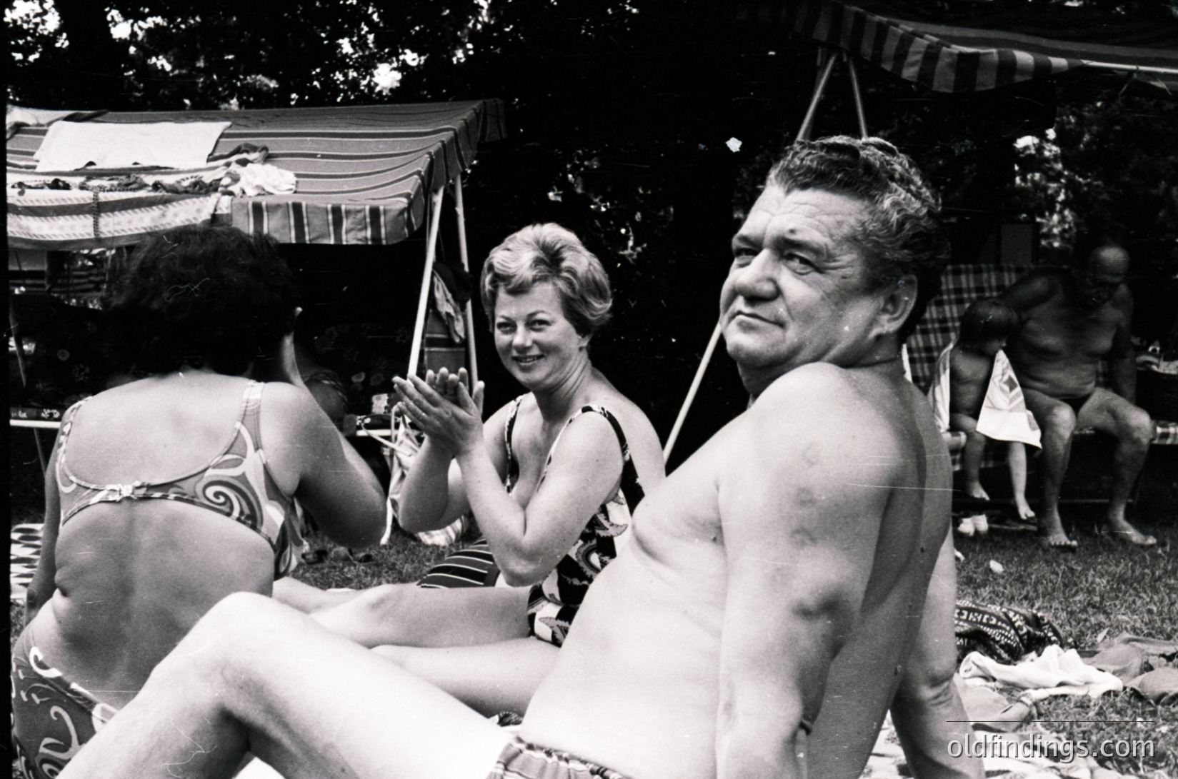 A 1960s beach scene captures three adults under a striped canopy, likely at a resort. The woman on left wears a patterned swimsuit with floral trim, while the man on right, shirtless, sits beside a woman in a sleeveless dress. Background shows more beachgoers and a boat docked near trees.