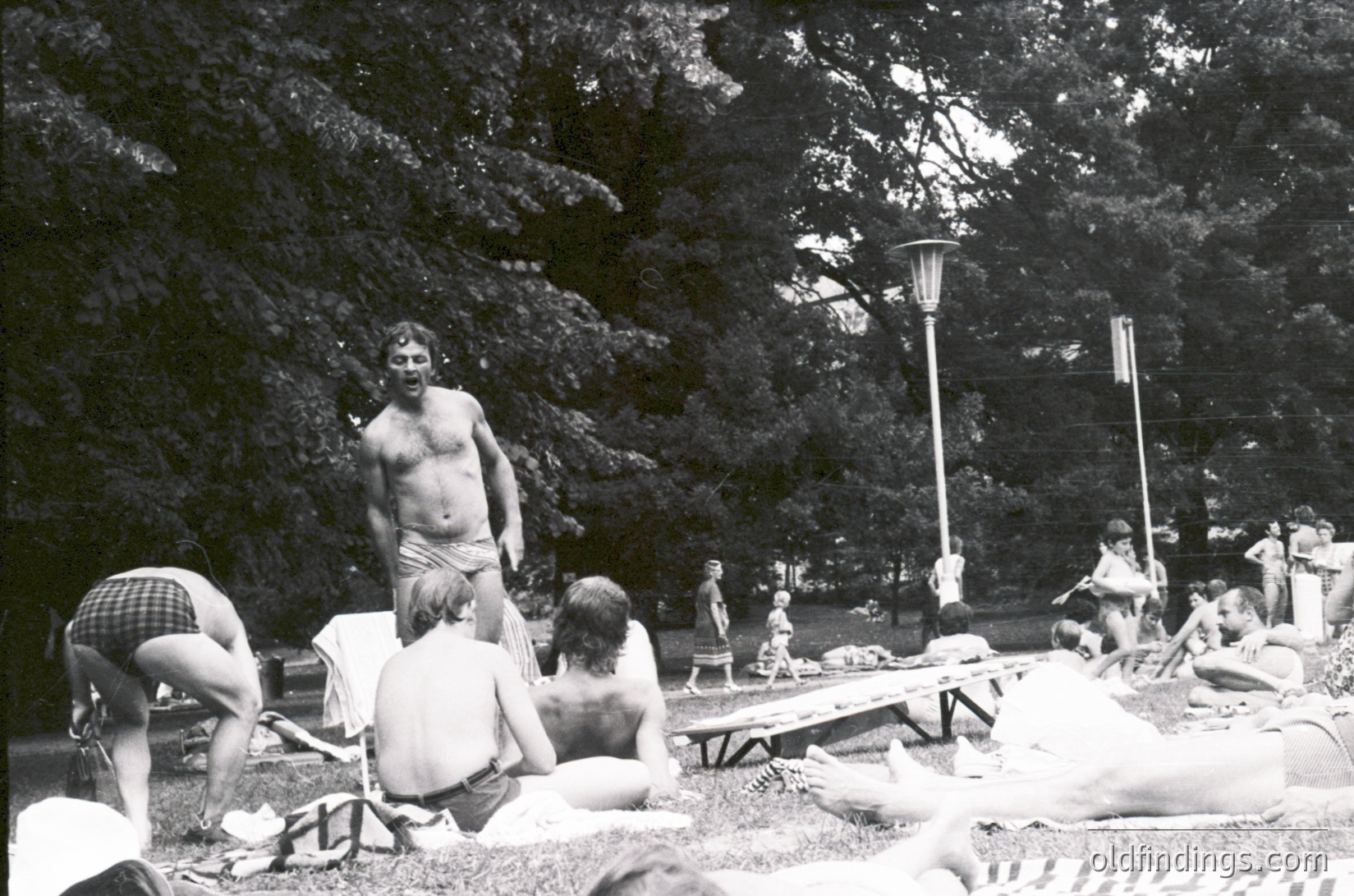 Black-and-white snapshot of a mid-20th-century outdoor gathering in a wooded park. Central figure stands shirtless, surrounded by relaxed groups on blankets and benches. Classic 1960s-70s attire—plaid shirts, shorts, and sandals—evokes a carefree era. Lamp post and picnic tables suggest a designated public space.
