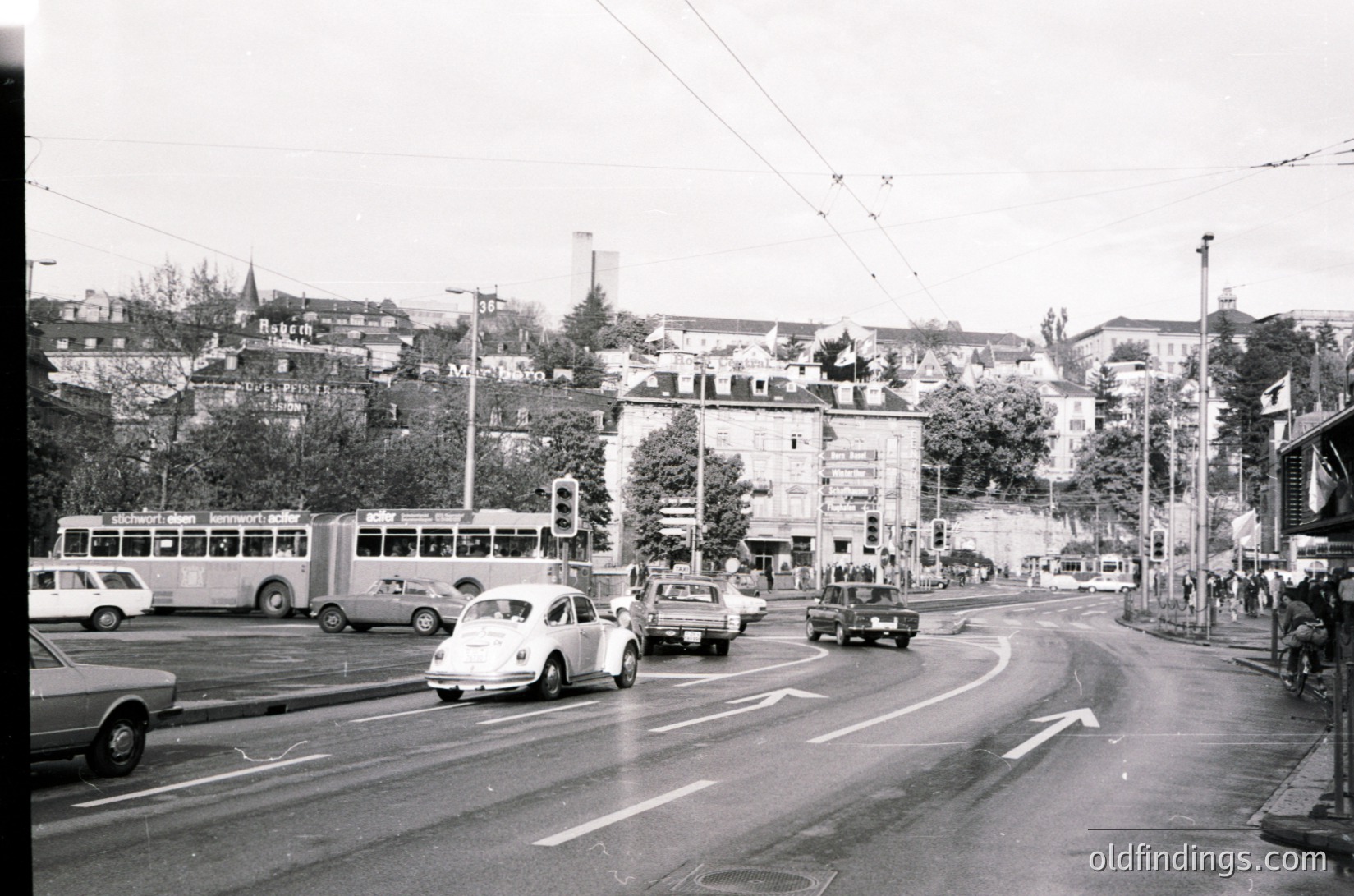 Mid-20th century urban intersection with vintage vehicles, including a classic bus and sedans, navigating a curved road. Overhead trolley wires and traffic lights indicate a mixed transit system. Dense residential buildings and greenery on a hillside suggest a European cityscape, likely --- *Note: Specific location cannot be confirmed without clearer landmarks, but the style aligns with mid-20th-century European urban design.*