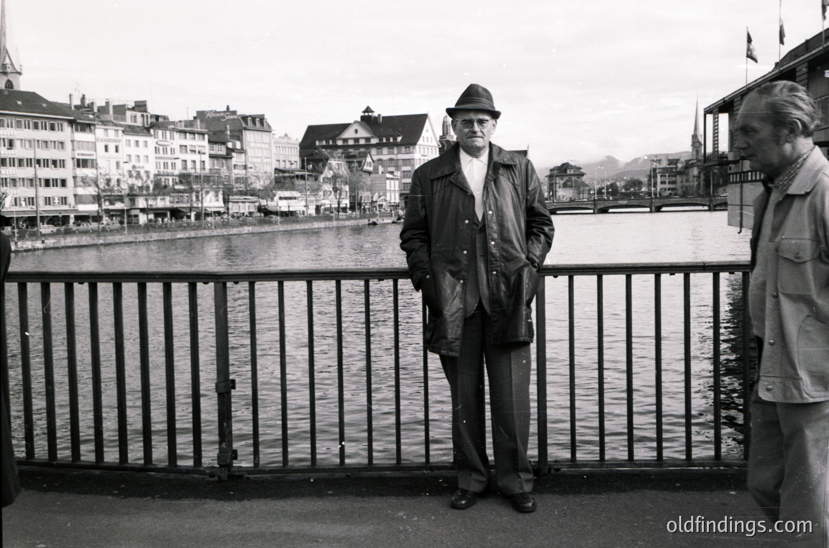 Mid-20th century man in fedora and leather jacket poses by a lakeside railing, with European-style buildings and waterfront in background. Likely , Switzerland, .
