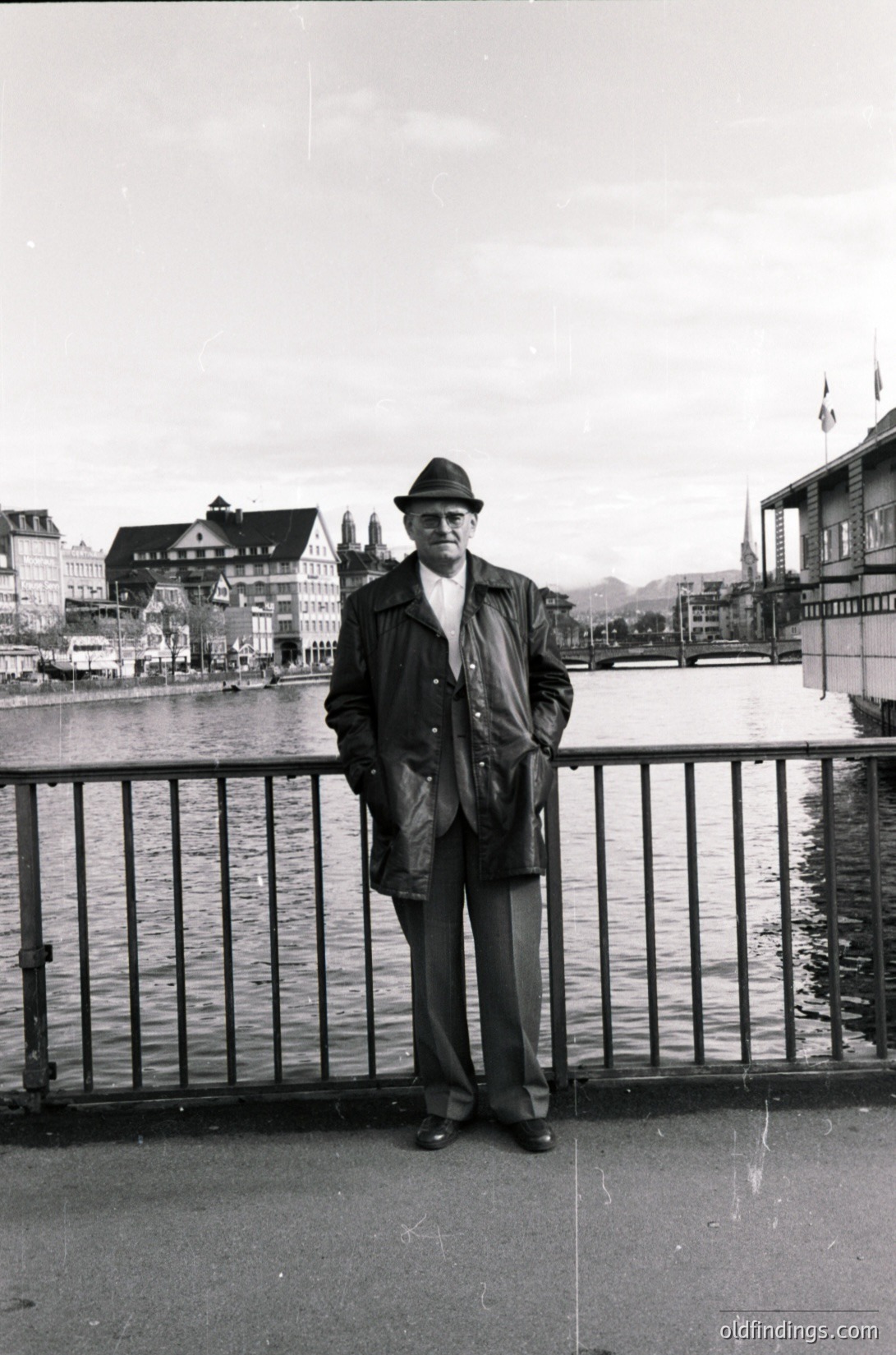 Mid-century man in a fedora and trench coat poses by a waterfront bridge, likely Zurich’s Lake Zurich. Architectural details include Art Deco-style buildings and a distant church spire. Black-and-white, 1950s–1960s.