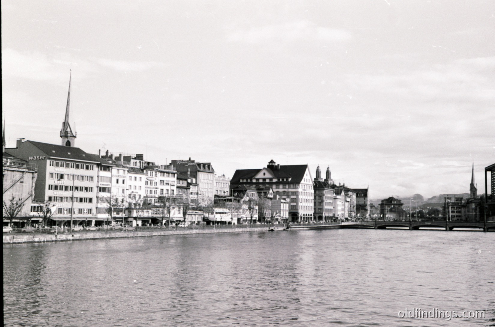 Black-and-white waterfront view of Zurich’s Limmat River, showcasing early-to-mid 20th-century European architecture. Prominent buildings feature gabled facades, shuttered windows, and a mix of residential/commercial use. A church spire and distant mountains frame the scene.