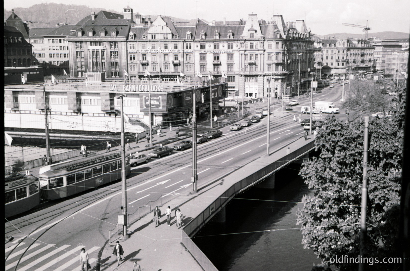 Vintage black-and-white urban scene featuring a tram crossing a bridge over a river in a European city. Mid-20th century architecture with multi-story residential and commercial buildings lining both sides. Prominent signage on buildings suggests retail or hospitality use. Light traffic, including cars and pedestrians, indicates active city life.