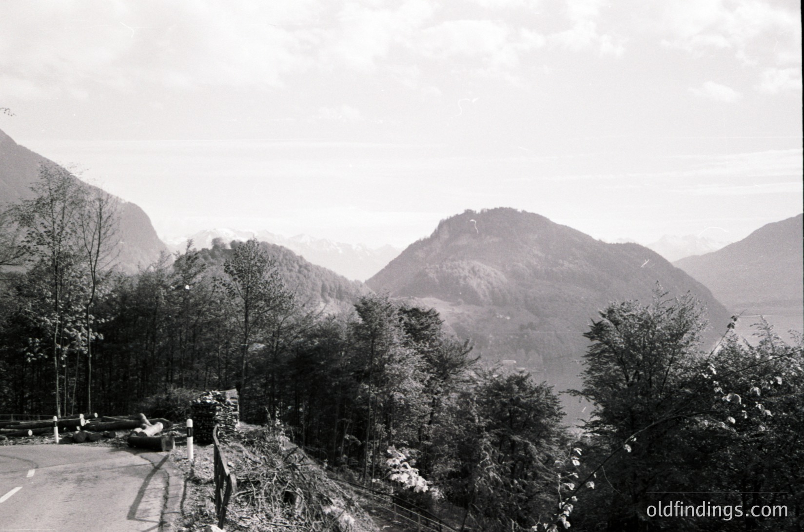 Black-and-white mountain landscape featuring rugged peaks and sparse forest. Road curves along base with minimal traffic. Overcast sky enhances dramatic, timeless atmosphere. Likely mid-20th century due to monochrome and styling.