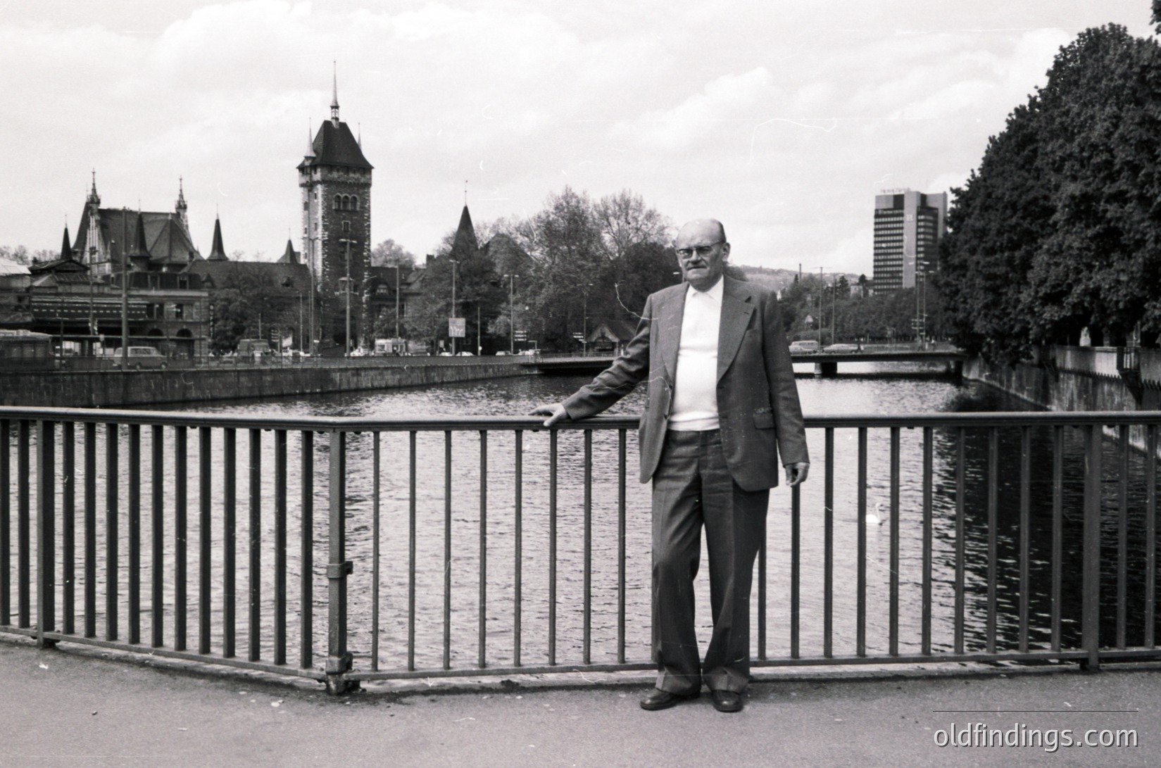 Mid-20th century black-and-white portrait of an elderly man in a suit, leaning on a metal bridge railing beside a river. Historic European architecture—turrets, spires, and a clock tower—visible in background. Likely