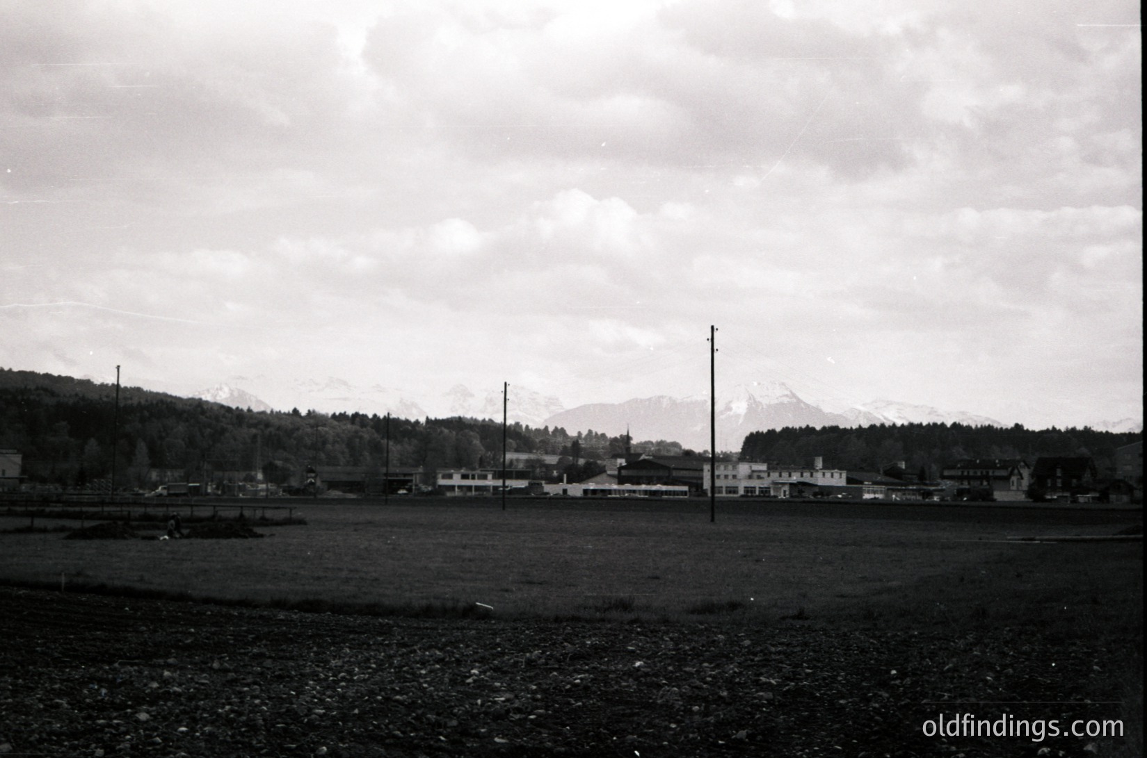 Black-and-white landscape featuring a flat, open field with sparse vegetation, likely agricultural. Distant village with low-rise buildings and a single prominent tower. Snow-capped mountains frame the horizon under overcast skies. Mid-20th century rural European setting.