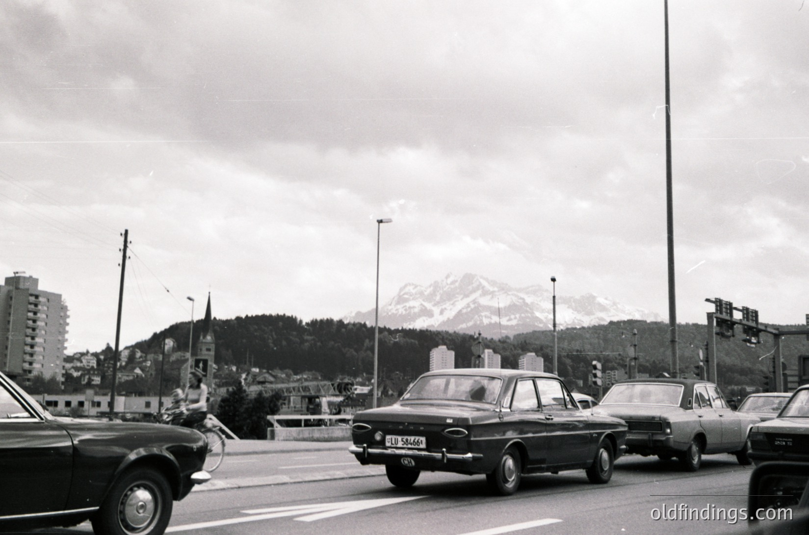 Mid-20th century urban traffic scene with vintage cars on a multi-lane road. Snow-capped mountain range and dense forest in background, suggesting alpine city. Mid-rise residential buildings and church spire visible. Overcast sky enhances vintage monochrome aesthetic.