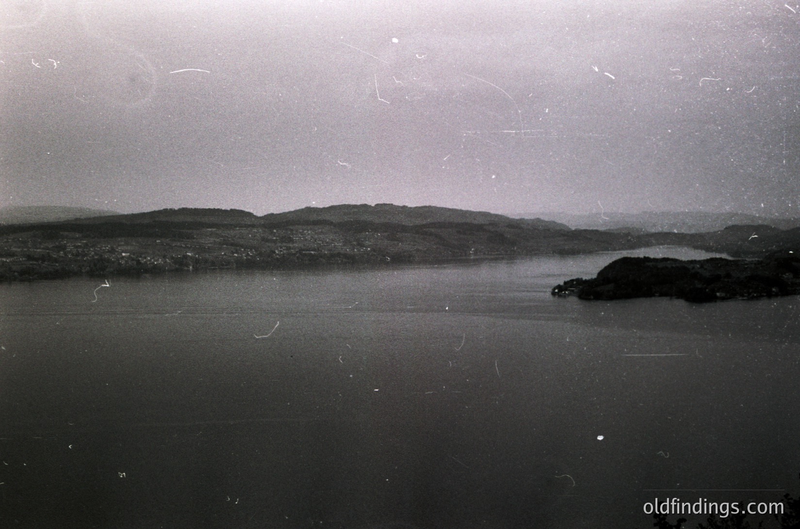 Vintage black-and-white coastal landscape featuring calm waters and distant hilly terrain. Distinctive fog or mist obscures mid-ground, enhancing depth. Possible or era due to grain and toning. Ideal for or .