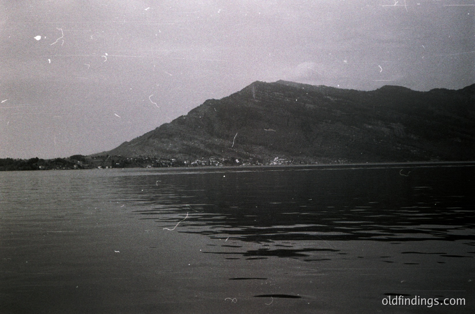 Vintage black-and-white coastal scene featuring jagged mountain range framing a calm body of water. Distinctive ripples and slight haze suggest early 20th-century photography. Coastal village nestled at base of mountains visible in background.