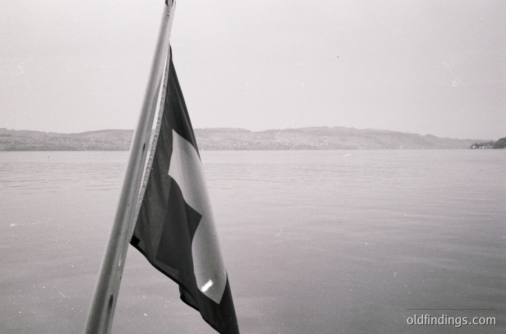 Black-and-white flag with red star and sickle on a pole, likely Soviet-era, over calm waters with distant hills. Mid-20th century maritime or coastal setting.