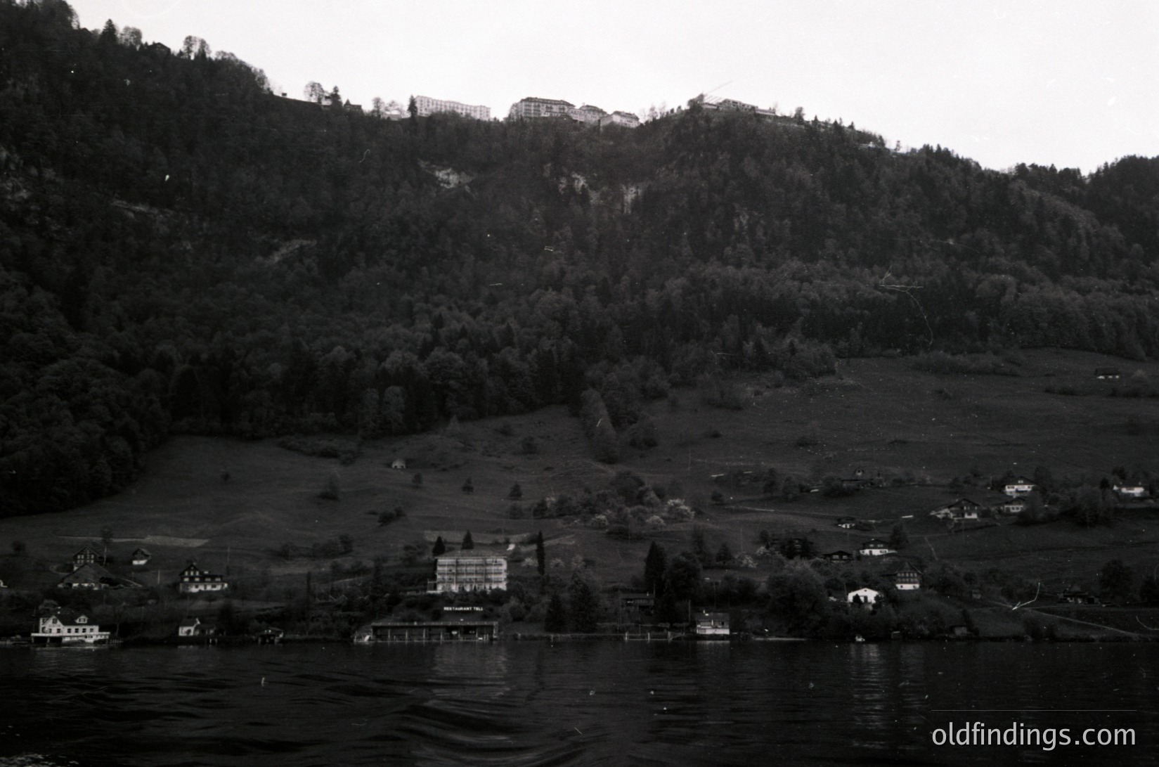 Black-and-white lakeside village nestled between forested hills and a rocky cliffside. Prominent lakeside resort with docks and scattered homes along the shoreline. Dramatic mountain backdrop suggests alpine or highland geography. Timeless composition ideal for travel, architecture, and nature photography.