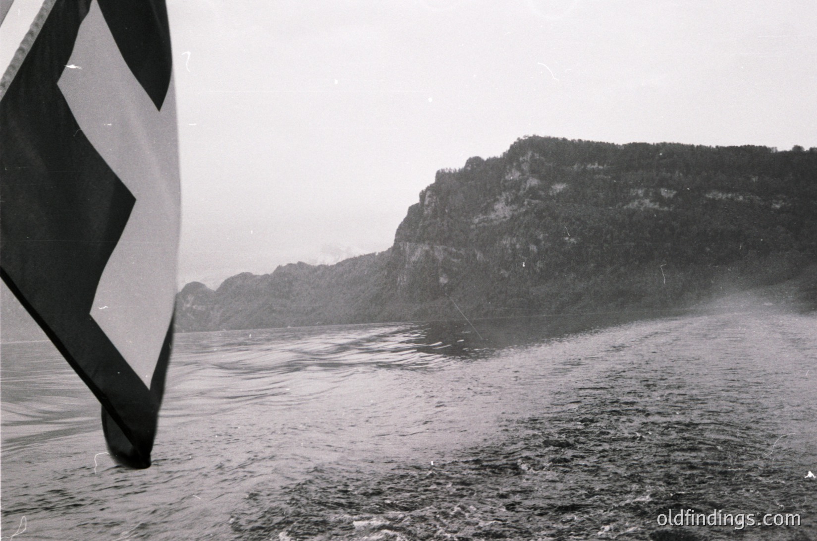 Black-and-white shot of a sailboat’s hull and sail against rugged cliffs, likely a fjord or coastal inlet. Dense forest covers steep rock faces, with mist or rain adding texture to the water. Composition suggests mid-20th century maritime photography, possibly or regions.