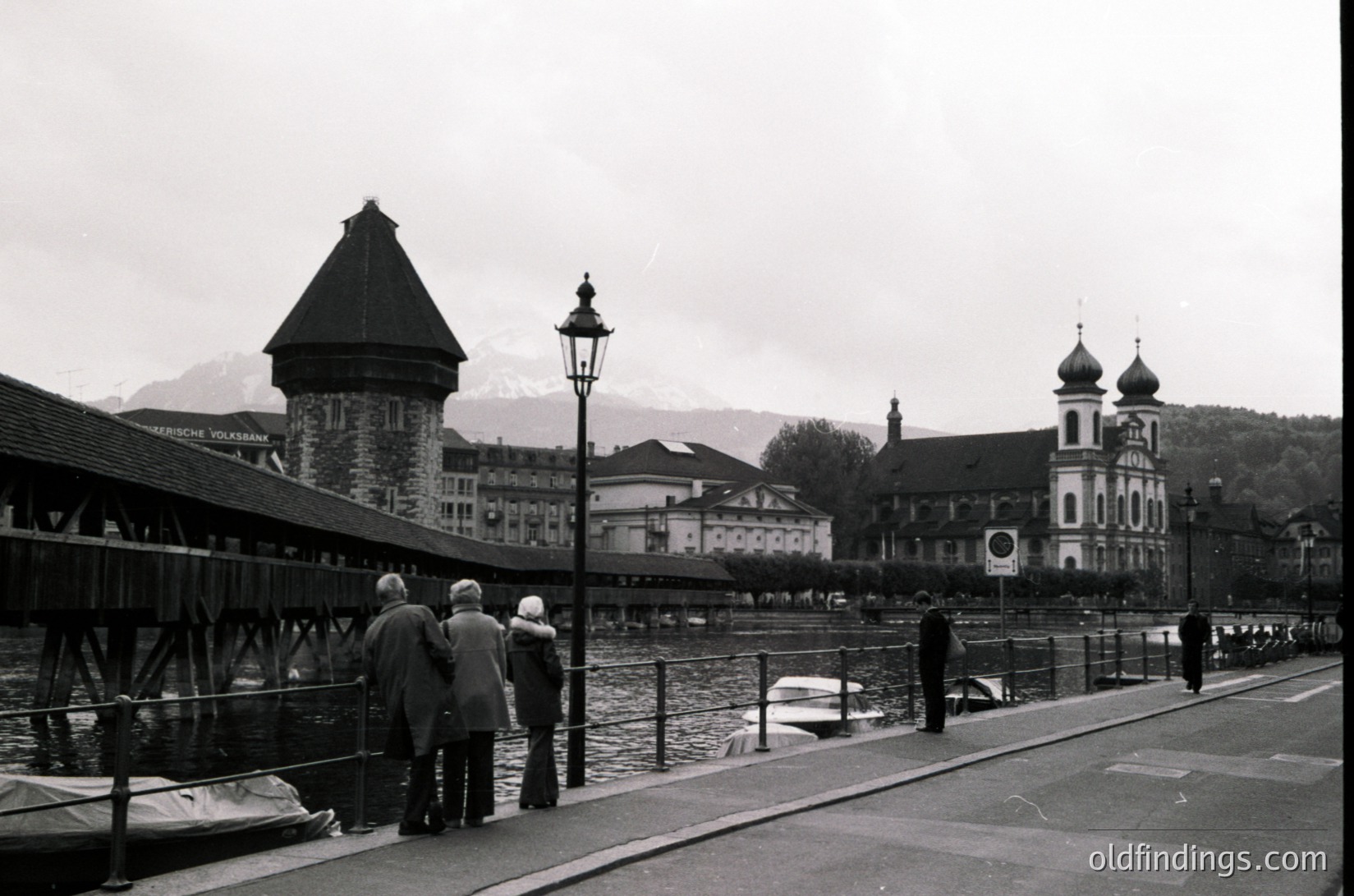 Black-and-white shot of the **Kapellbrücke (Chapel Bridge) in Lucerne, Switzerland**, featuring its iconic wooden bridge with 14th-century bell tower. Snow-capped Alps in background. Mid-20th century (1950s–60s) street scene with pedestrians and vintage streetlamps. ücke
