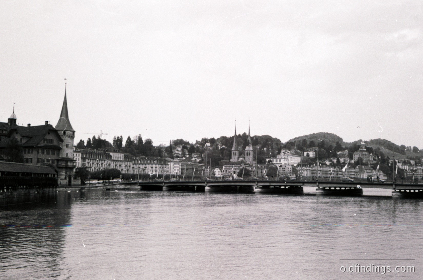 Black-and-white riverside cityscape featuring historic European architecture. Prominent spires, gabled roofs, and a bridge span the water, suggesting a mid-20th century urban setting. The calm river reflects muted city lights under an overcast sky.