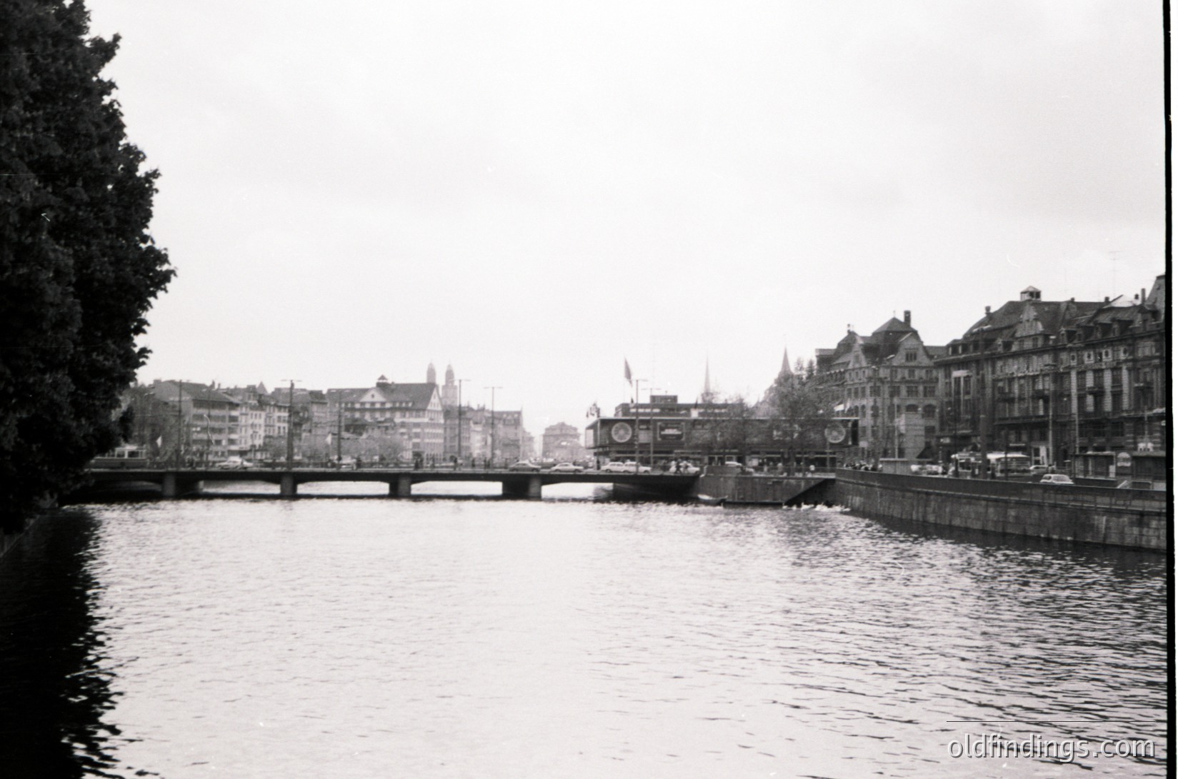 Mid-20th century urban riverscape featuring a stone bridge spanning a calm waterway. Historic European architecture with gabled facades and a prominent clock tower in background. Overcast skies and minimal traffic suggest a quiet moment. Likely