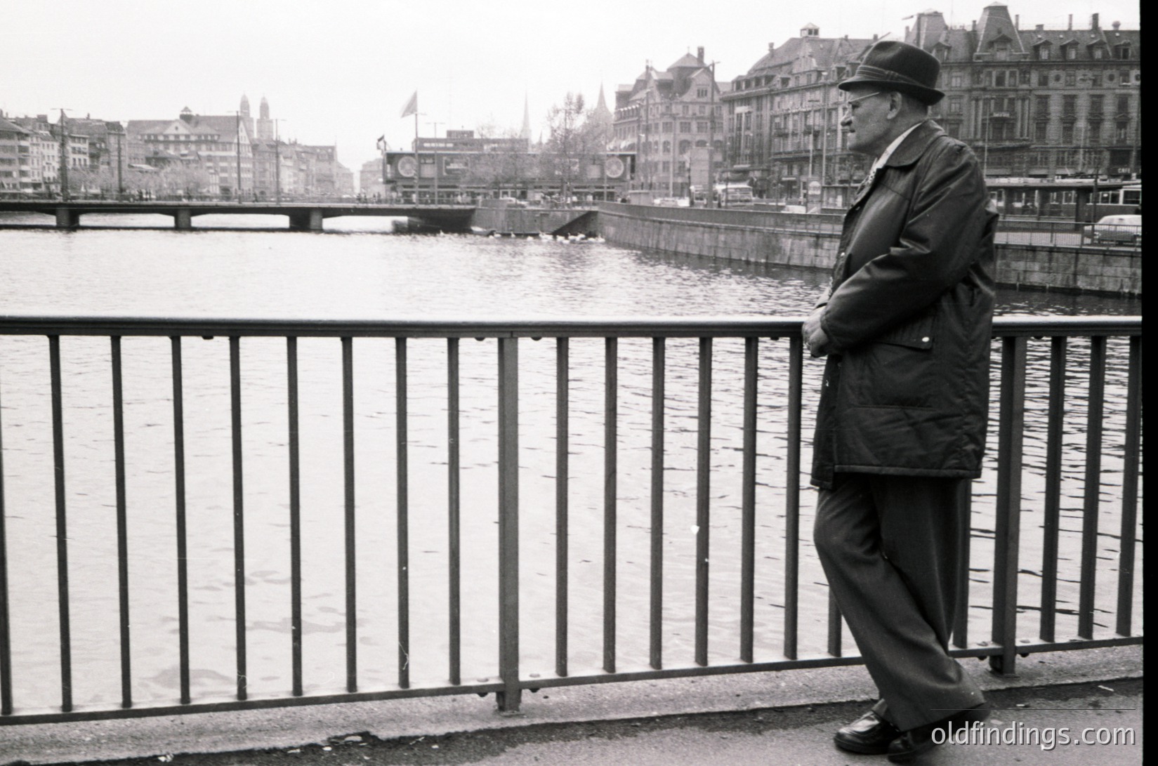 Mid-century man in a fedora and overcoat stands contemplatively on a bridge railing, overlooking a river in a European city. Historic architecture and a bridge with a flagpole in background suggest