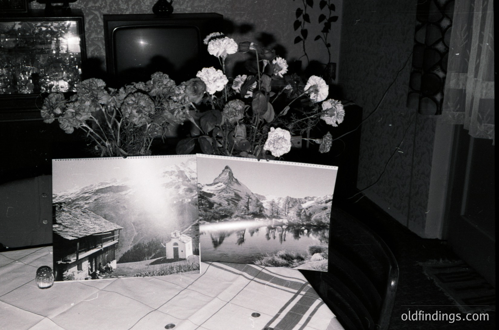Vintage indoor scene featuring a mid-century living space. Two framed black-and-white postcards depict alpine scenery—one showing a cabin by a lake, the other a mountain reflection. A bouquet of dried flowers sits on a tiled table beside a CRT TV and wooden cabinet. Decor suggests 1970s-1980s European home decor.