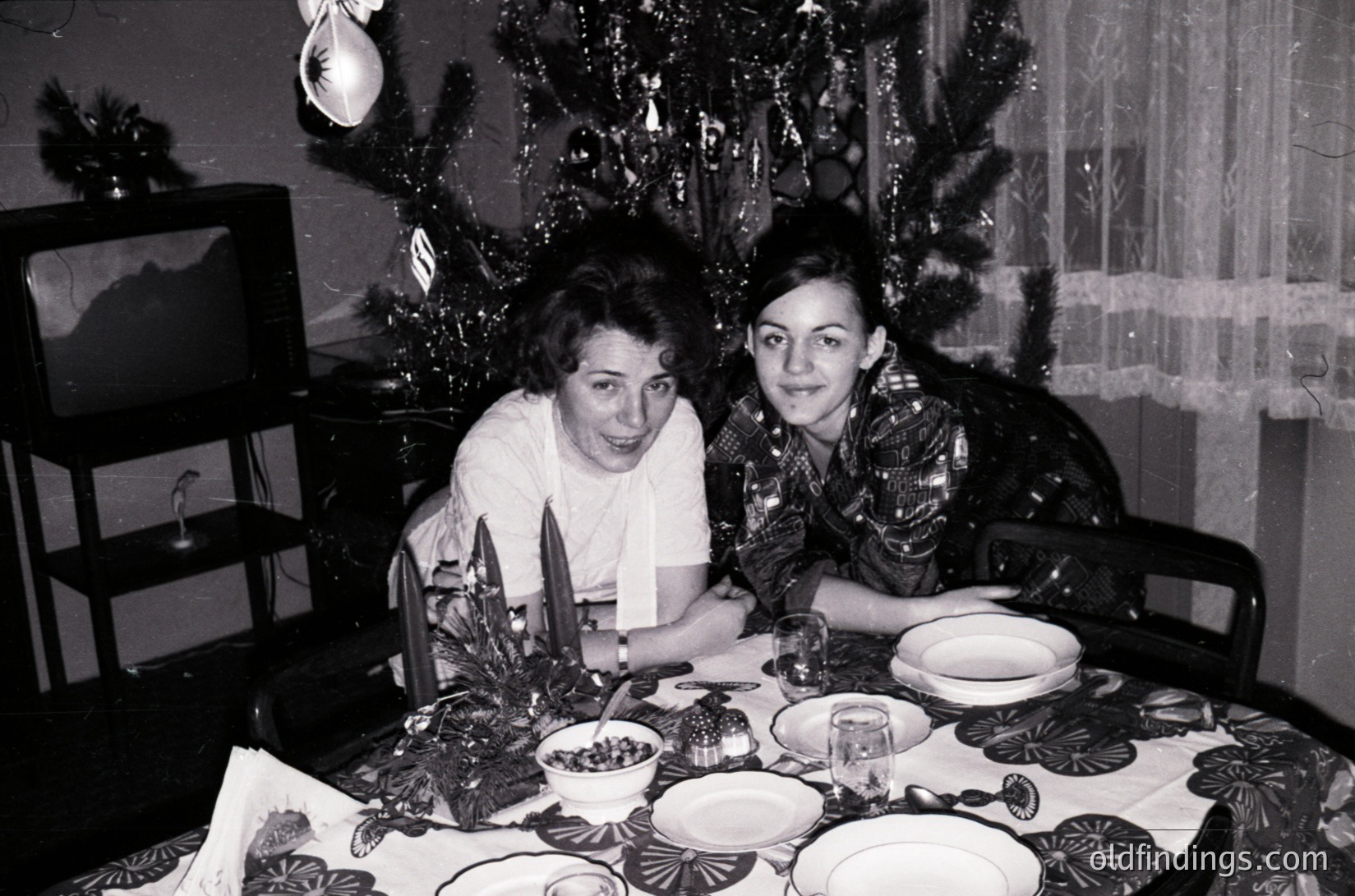 Two women pose at a festively set table in a mid-century home, likely 1950s–1960s. Decor includes a decorated Christmas tree, ornate plates, and vintage glassware. The setting reflects post-war European holiday traditions.