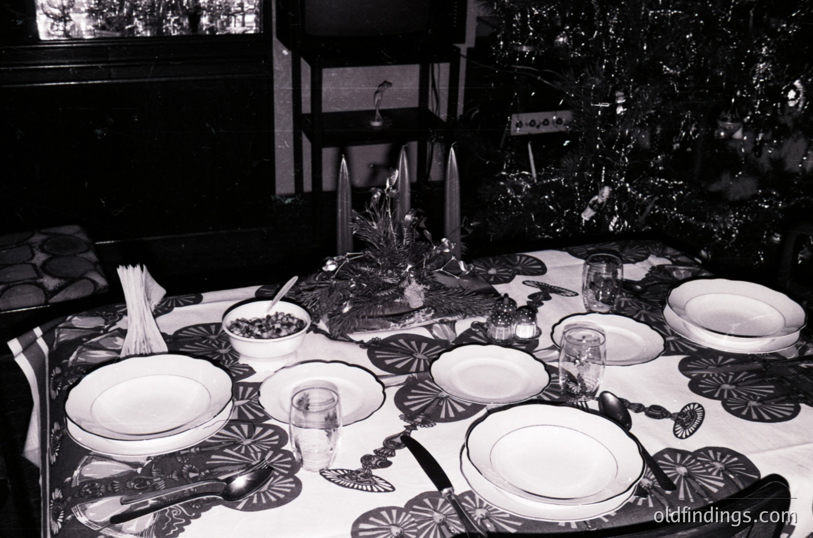 Elegant 1950s-60s holiday dining setup with symmetrical tableware: white plates, silverware, and glassware on a dark tablecloth with geometric patterns. Centerpiece features a tiered stand with decorative items and candles. Christmas tree visible in background, suggesting festive decor.
