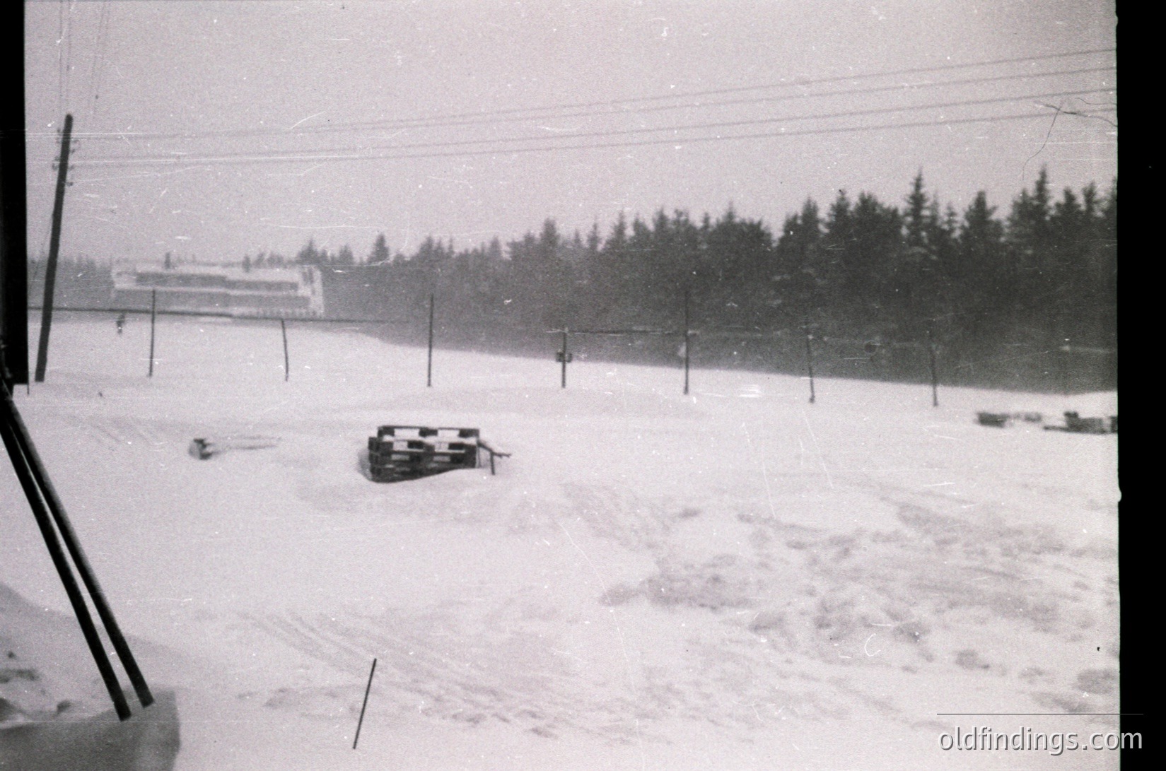 Snow-covered alpine slope with vintage sleds, likely mid-20th century. Dense forest and a multi-story building in background. Heavy snowfall obscures visibility.