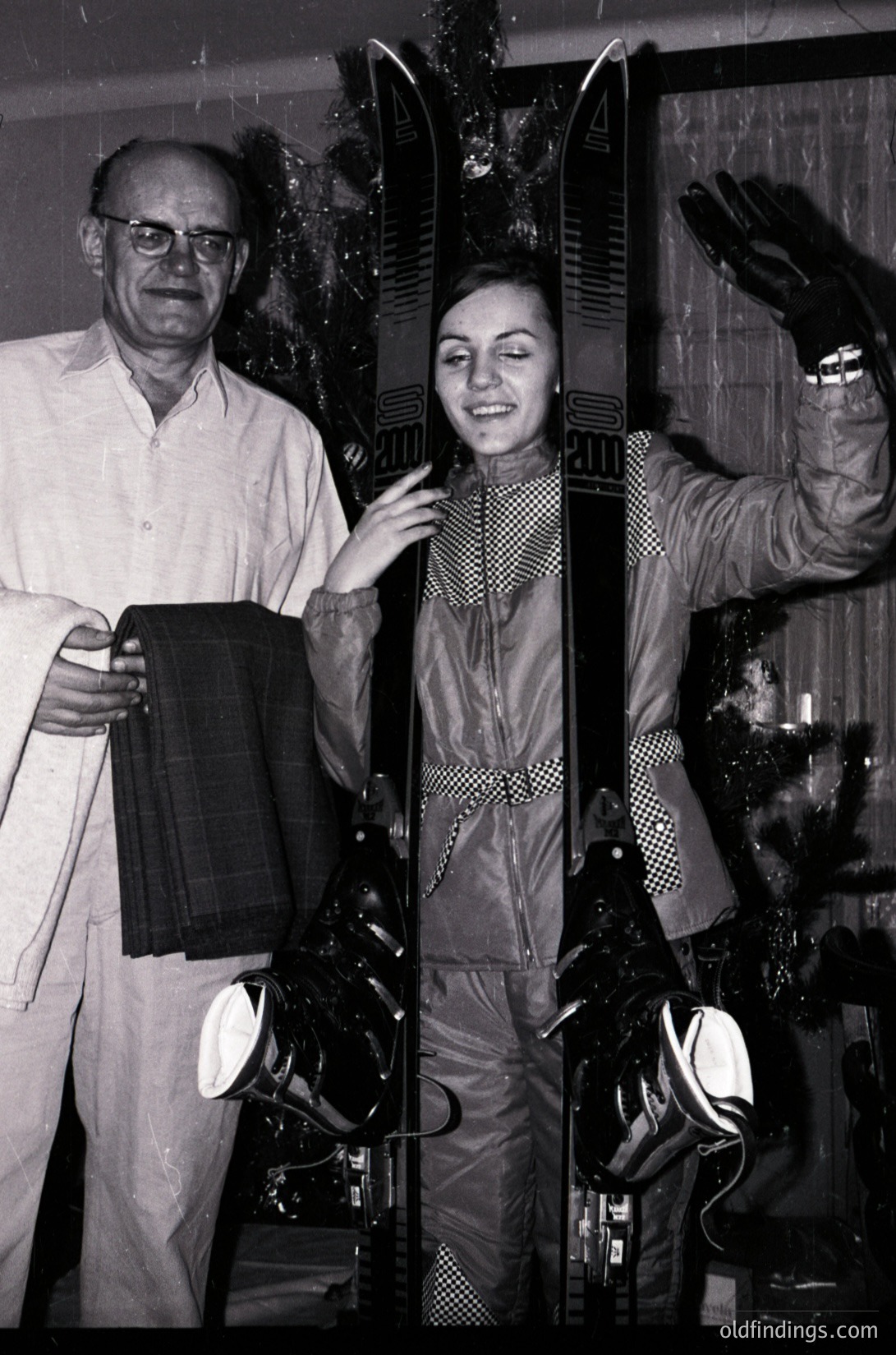 A man and woman pose indoors with alpine gear, likely in a ski shop or training facility. The man holds a pair of skis while the woman wears patterned ski attire and gloves, holding ski poles. The setting appears to be mid-20th century, possibly the 1960s–1970s, with vintage ski equipment and lighting. [Vintage ski shop portrait with man and woman in alpine gear, 1960s–1970s ]