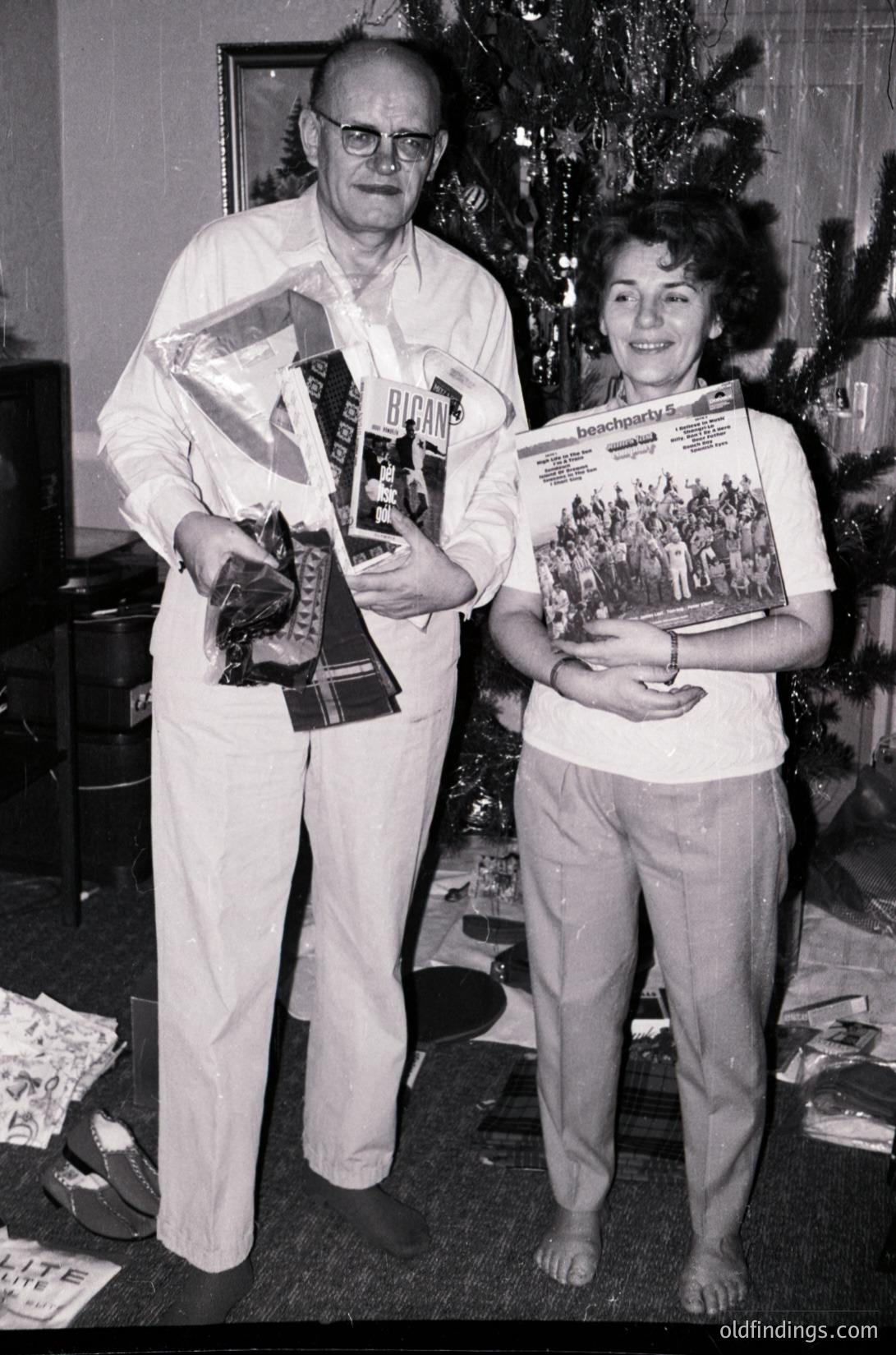 A man and woman pose indoors beside a decorated Christmas tree, holding awards and magazines. The man wears glasses, a white robe, and holds a trophy and a *Bulgarian* magazine. The woman holds a *Descendants* magazine. The setting suggests a festive 1970s-80s Bulgarian holiday celebration.