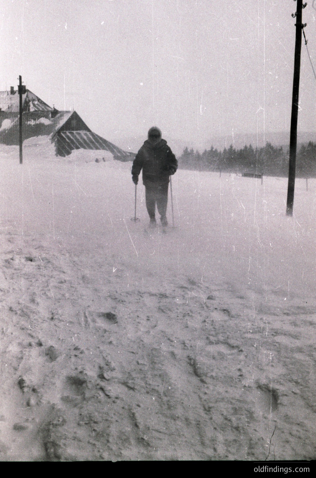 Black-and-white winter scene showing a lone cross-country skier in mid-motion on a snow-covered trail. Snow-laden trees and a wooden cabin in the background suggest a rural, alpine setting. Heavy snowfall obscures distant views. Likely mid-20th century (1940s–1960s) based on clothing and photographic style.