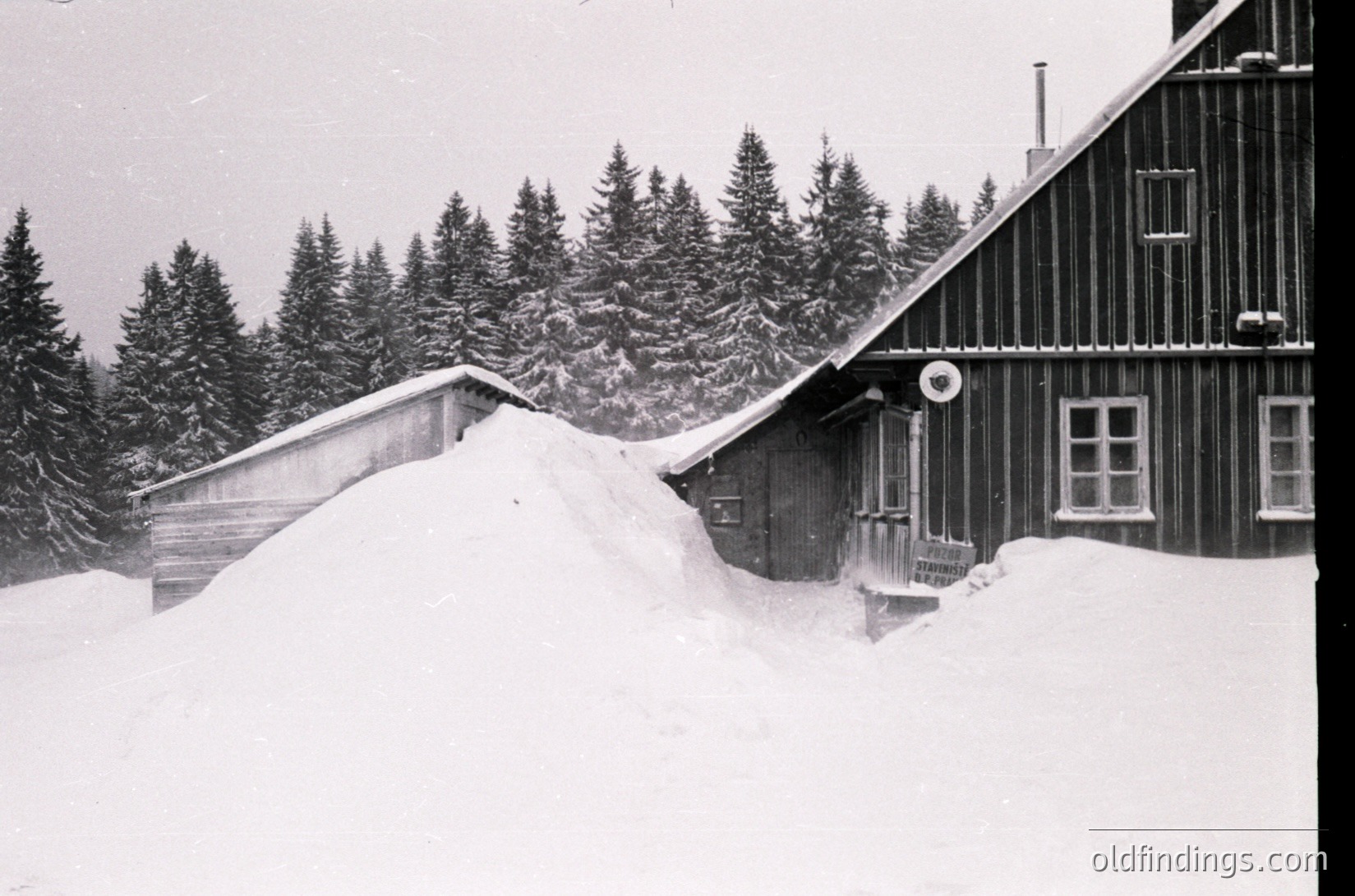 Mid-century alpine lodge buried under heavy snowdrift, showcasing classic wooden chalet architecture with sloped roof. Dense evergreen forest in background suggests high-altitude setting. Likely – European mountain resort.