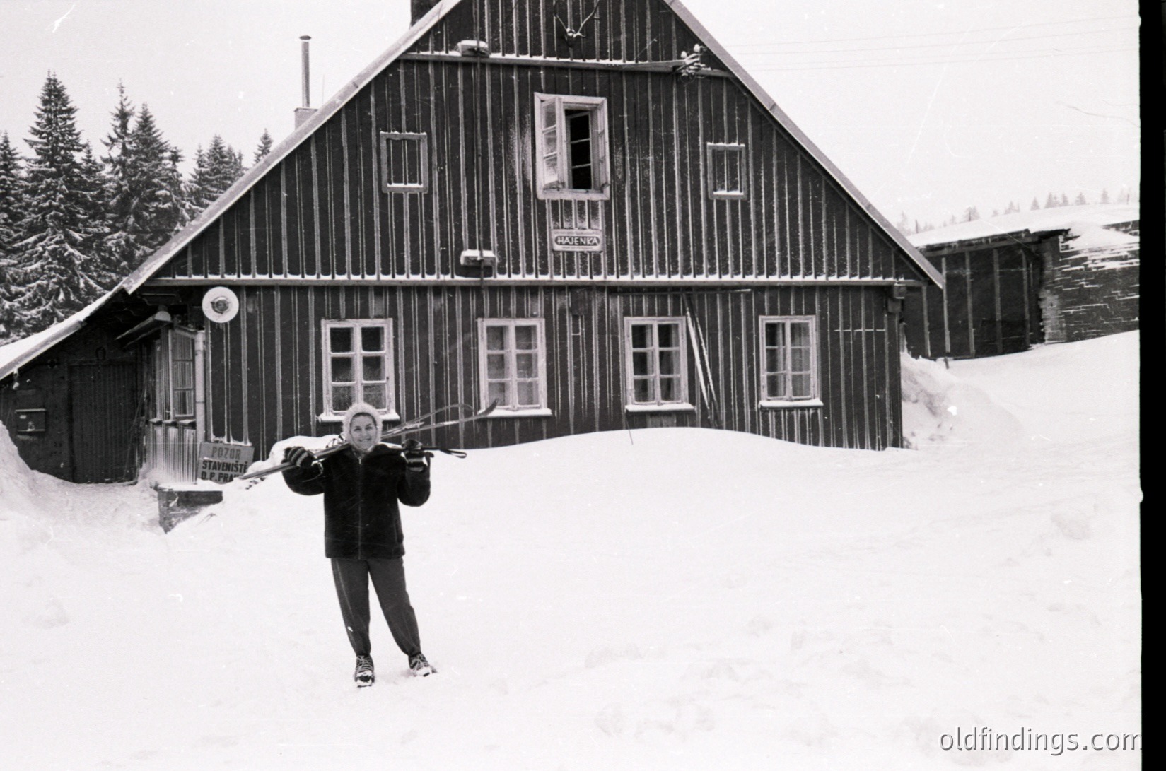 Black-and-white alpine lodge with wooden clapboard siding and white-framed windows, set in deep snow. A person in winter gear poses with skis, likely mid-20th century. Rustic mountain retreat with pine trees and snow-covered slopes.