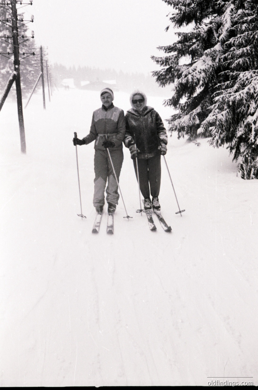 Two skiers pose on a groomed trail in mid-20th century alpine setting. Men’s and women’s ski attire from the 1950s-60s: padded jackets, knee-length skirts/pants, and classic wooden skis. Snow-laden evergreens frame the scene, hinting at European or North American winter sports culture.