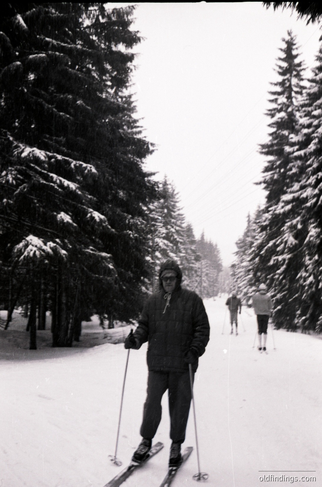 Mid-20th century cross-country skiing scene in a snow-covered forest. A man in vintage winter gear—wool hat, goggles, and a padded jacket—poses with ski poles on a groomed trail. Snow-laden evergreens frame the path, and another skier is visible in the distance. Likely or European alpine setting.