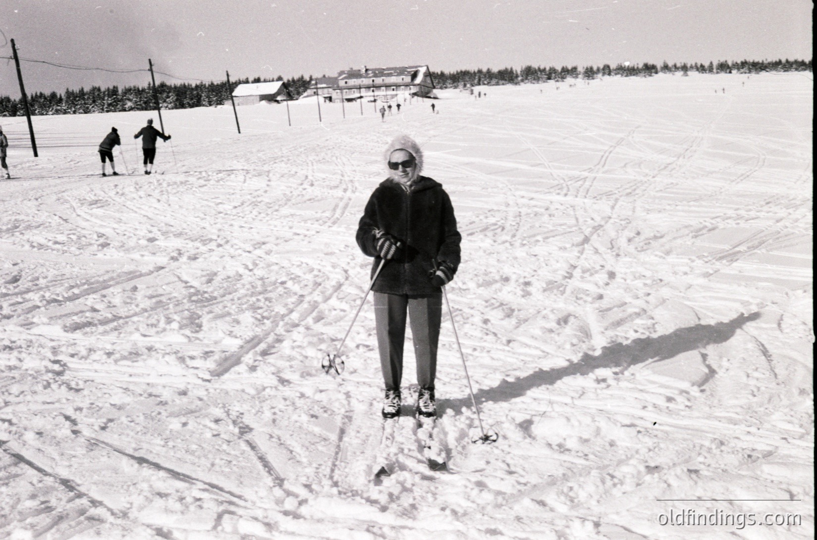 Mid-century cross-country skier pauses on groomed trail, wearing classic 1960s winter gear: knit hat, goggles, and long-sleeve top. Snow-covered forest and distant ski lodge in background suggest a Nordic skiing resort. Black-and-white captures vintage winter sports culture.