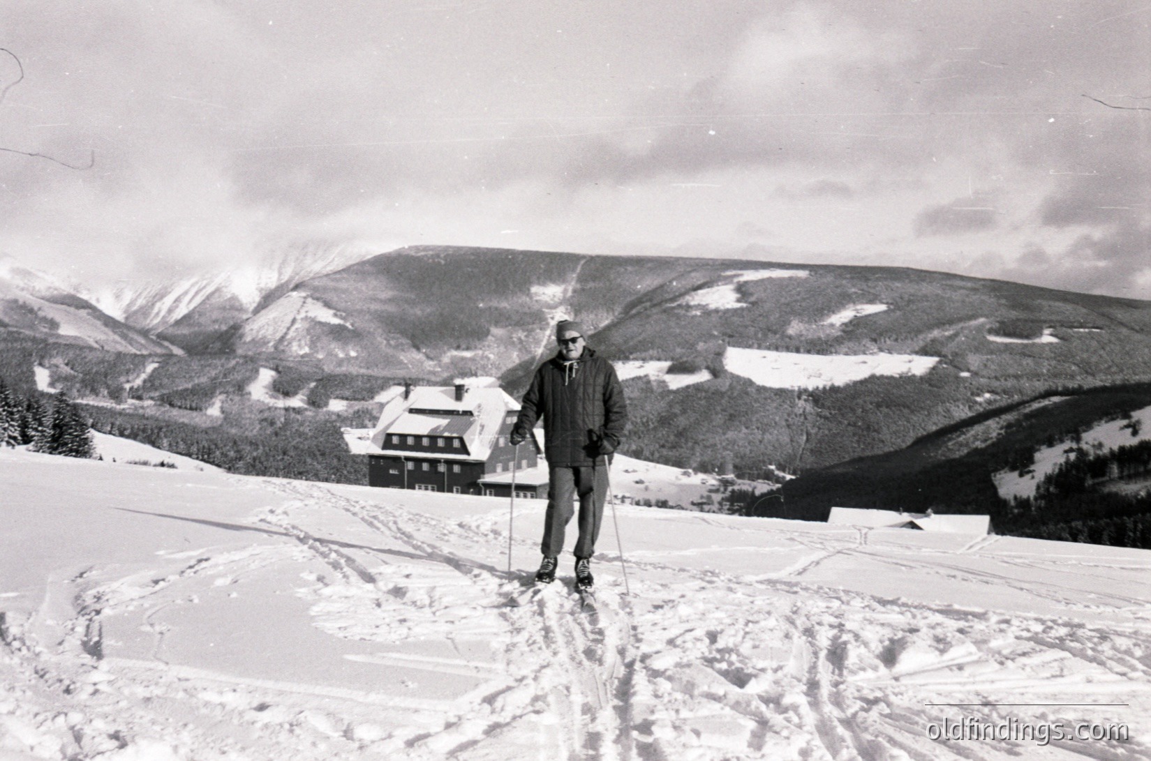 Black-and-white ski scene featuring a lone cross-country skier in mid-stride on groomed trails, surrounded by snow-covered alpine terrain. A rustic wooden chalet and forested slopes form the backdrop, suggesting a mid-20th-century European mountain resort.