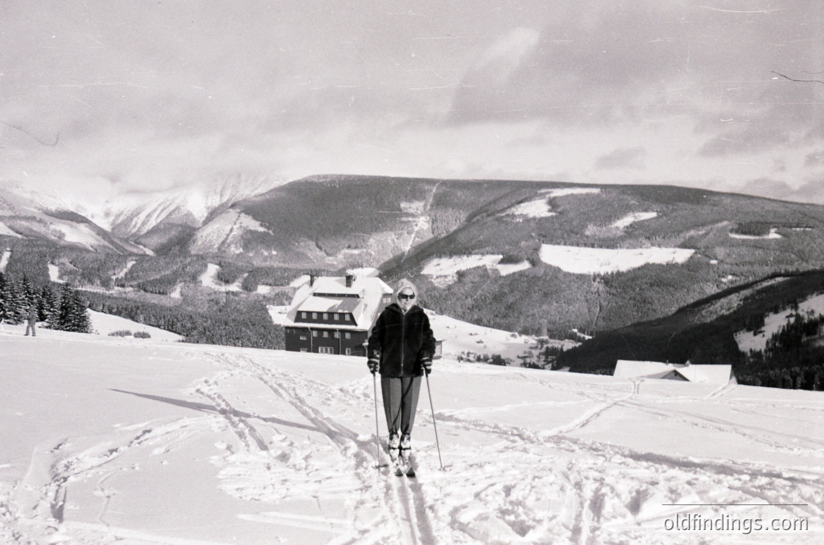 Mid-20th century cross-country skier posing on groomed trails in alpine setting. Wooden chalet-style lodge with sloped roof in background. Snow-covered valleys and forested ridges create dramatic backdrop. Classic 1950s-60s ski attire with goggles and long-sleeve shirt.