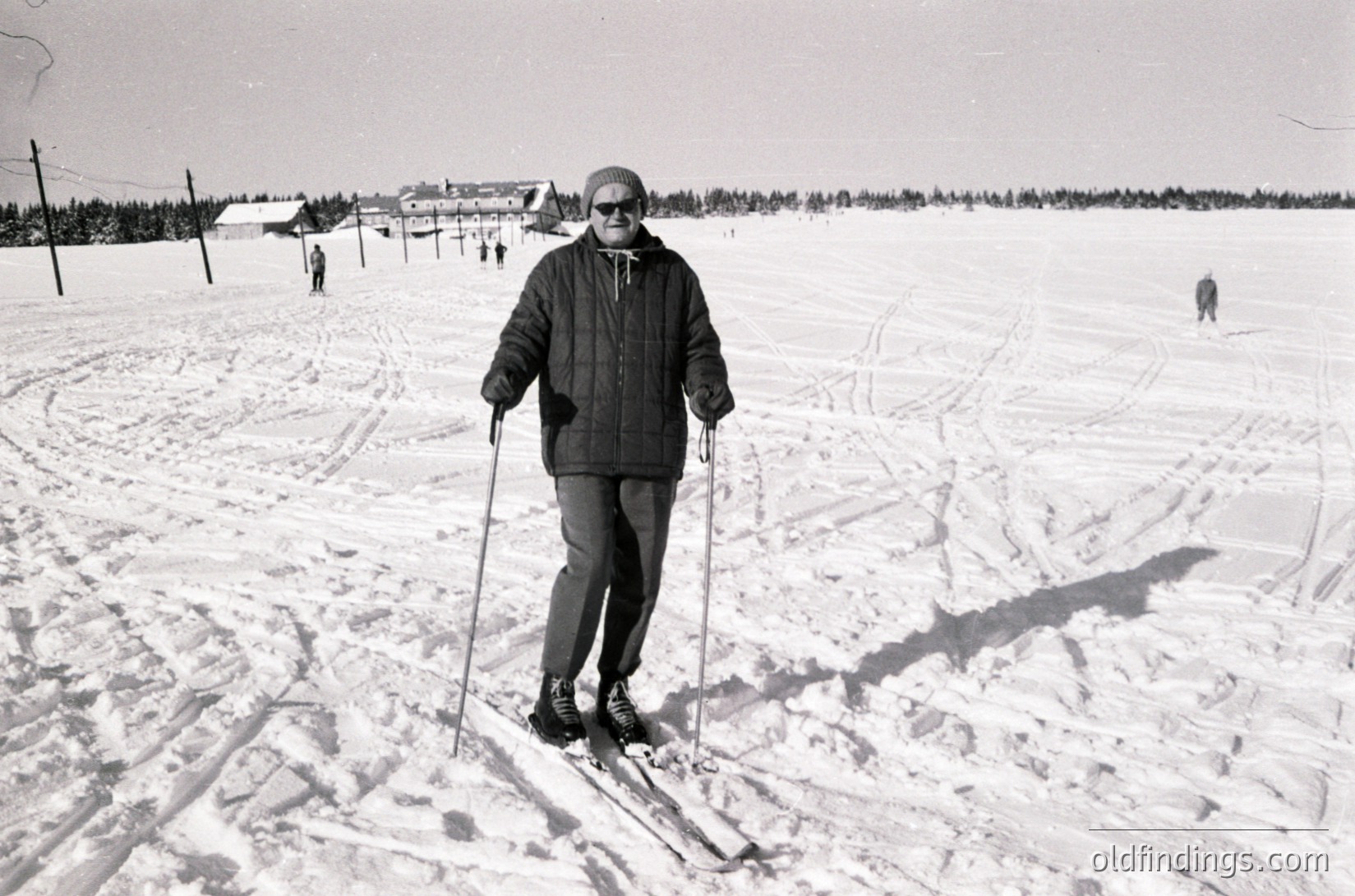 Mid-century cross-country skier in winter attire poses on groomed trail, surrounded by snow-covered landscape. Distant ski tracks and forested horizon suggest a Nordic skiing scene. Likely – era, or .