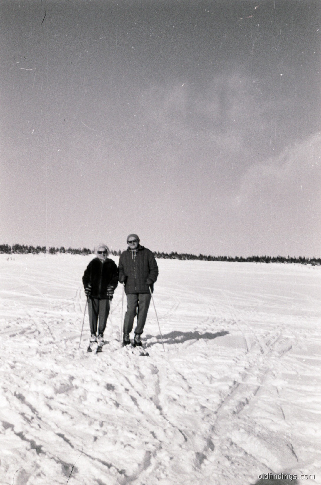 Mid-20th century cross-country skiing duo on snow-covered trail, likely or . Man in cap, woman in dark coat pose together, surrounded by forested horizon. Vintage or winter sports scene.